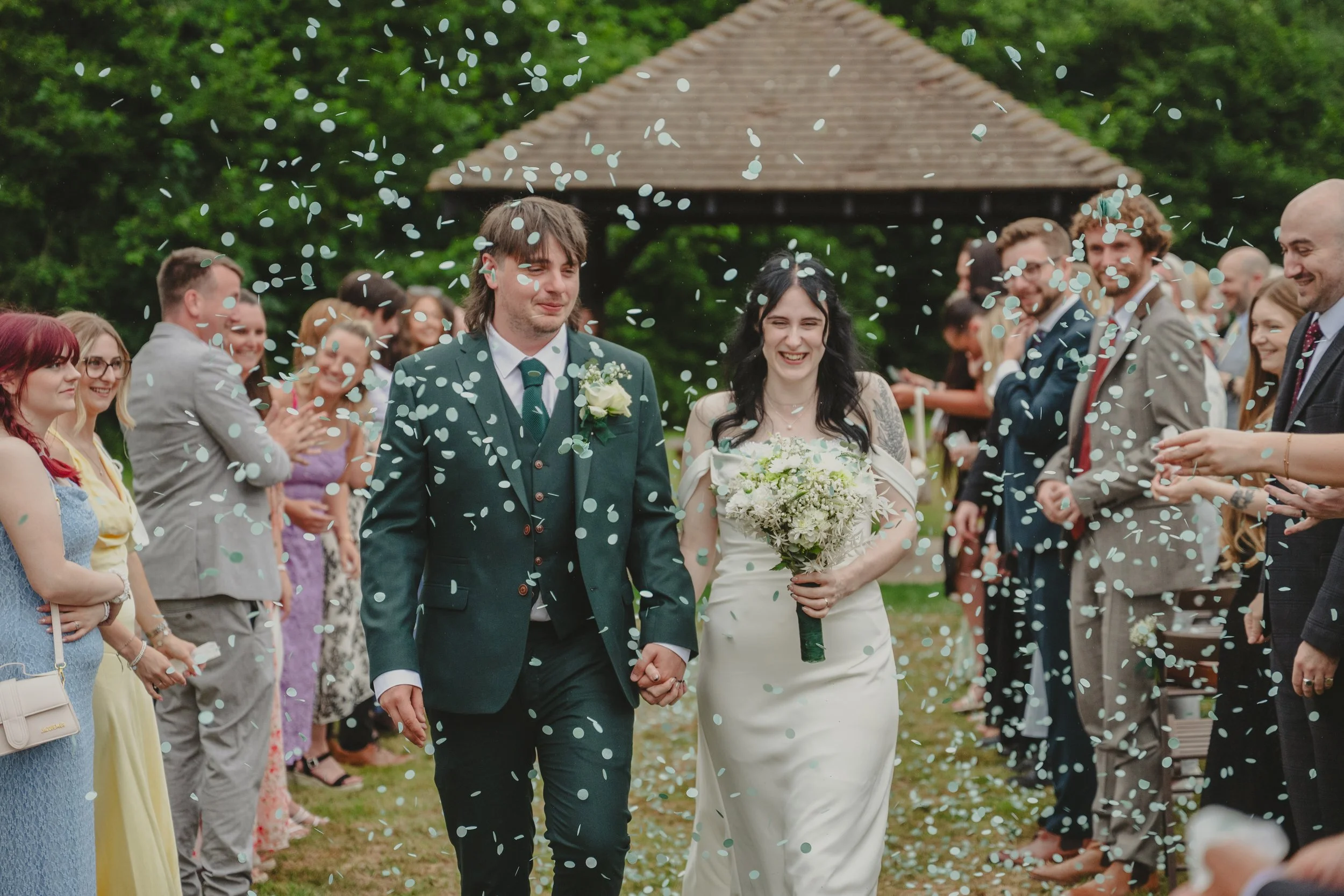A bride and groom holding hands and smiling at each other during their outdoor wedding as guests cheer and throw confetti.