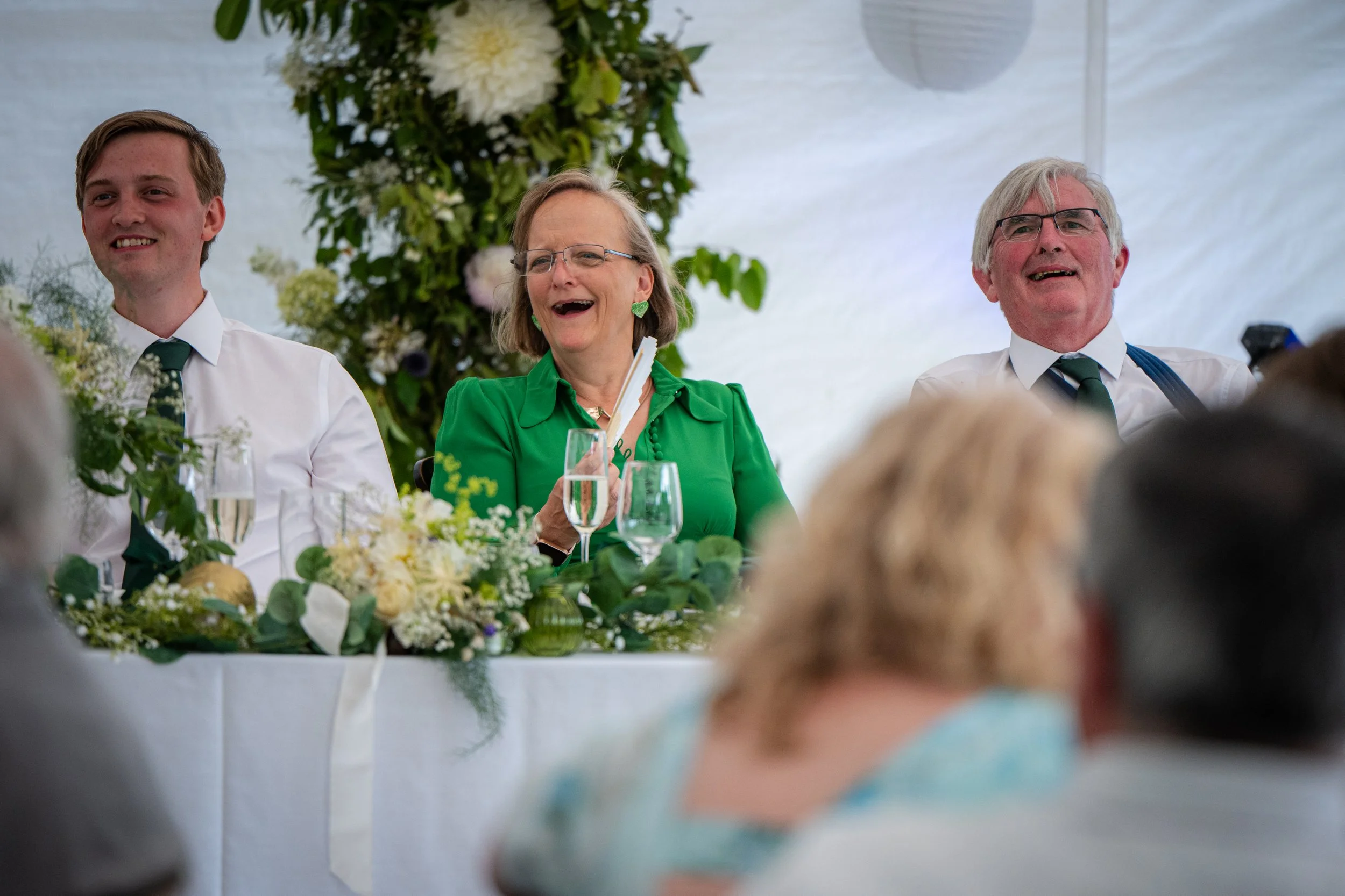 Three people sitting at a decorated table, smiling, with a floral arrangement behind them, in a bright setting.