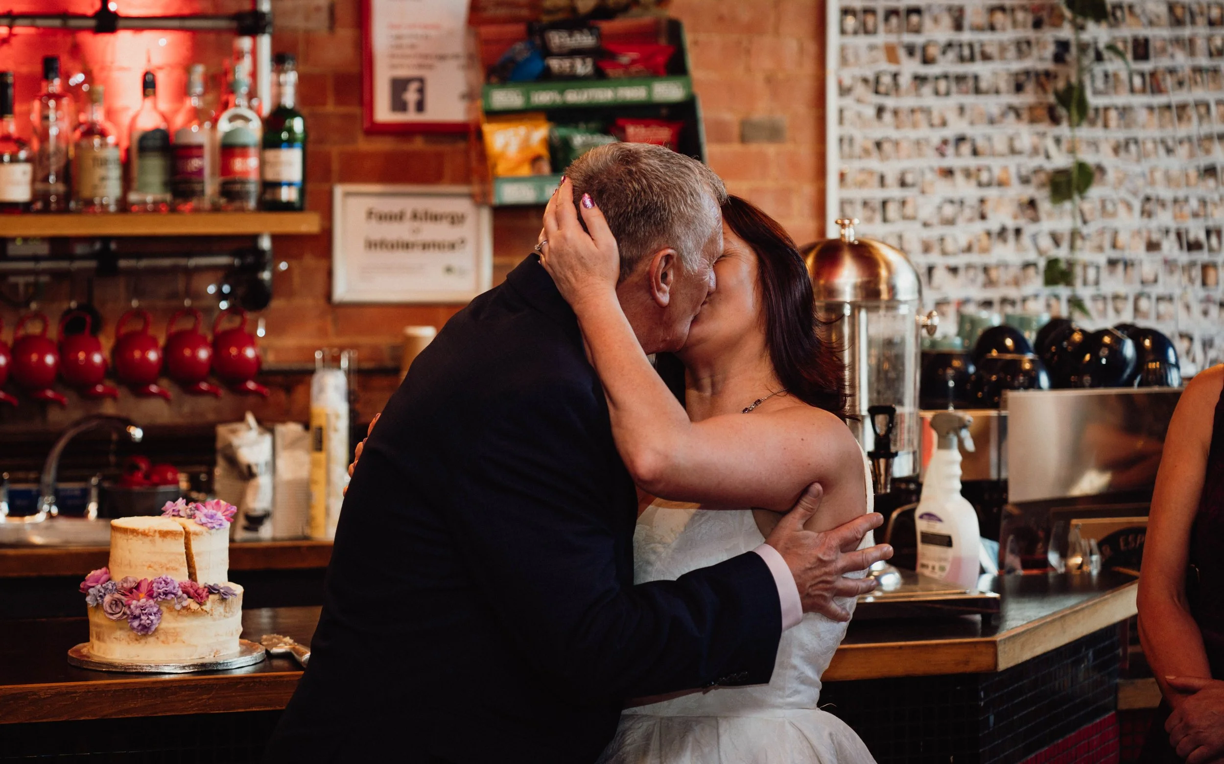 A couple sharing a kiss at a celebration in a cafe with a cake decorated with purple and pink flowers in the foreground.