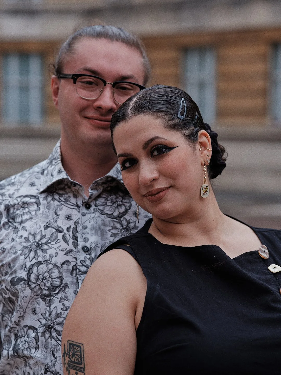 A young man and woman posing outdoors in front of wooden building. The man has glasses, wearing a floral shirt, with light skin and blonde hair tied back. The woman has dark hair with hair clips, wearing a black sleeveless top, with piercings, earrin