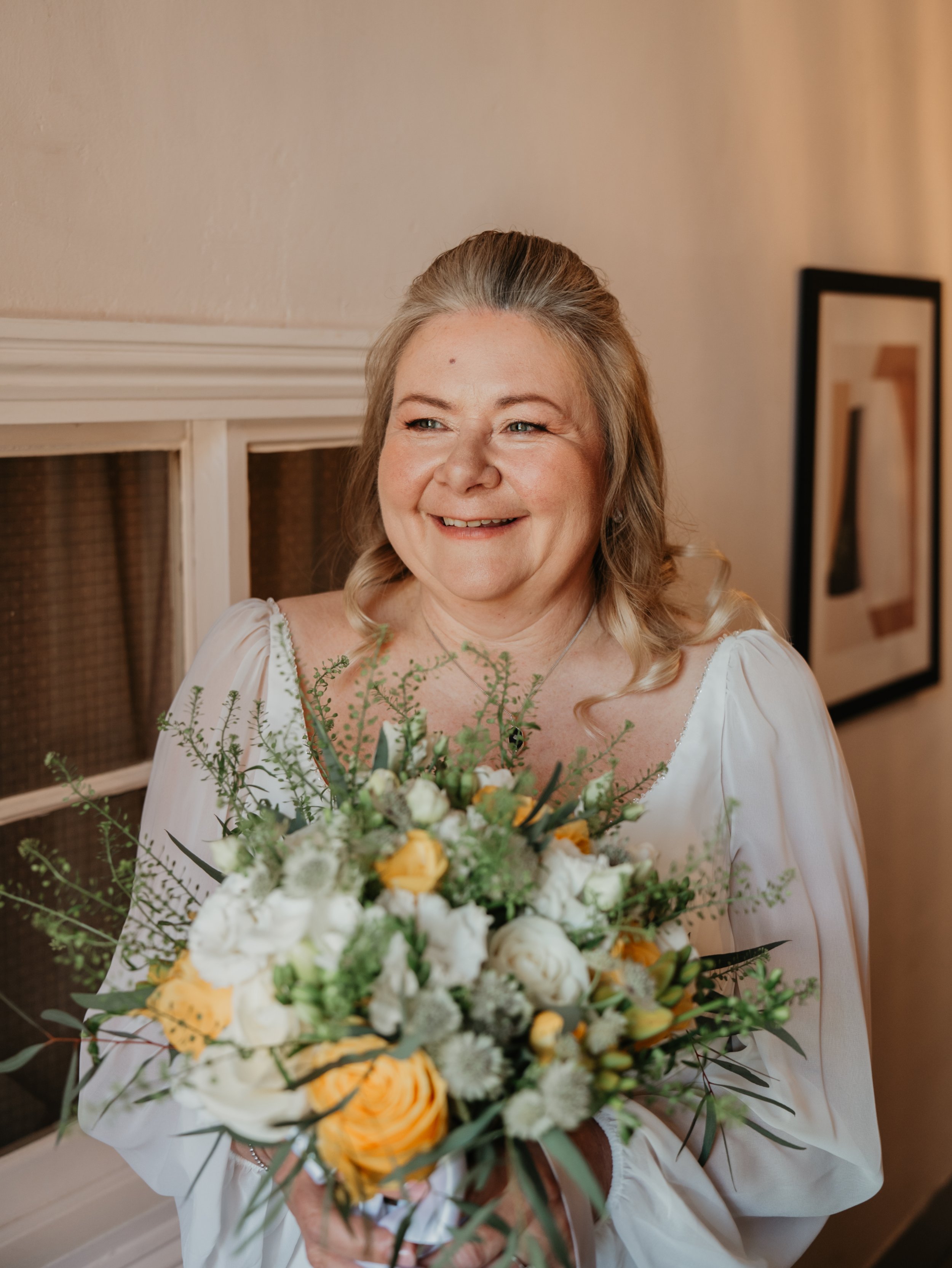A middle-aged woman smiling and holding a large bouquet of white, yellow, and orange flowers, wearing a white dress with lace details, standing indoors against a beige wall with framed artwork.