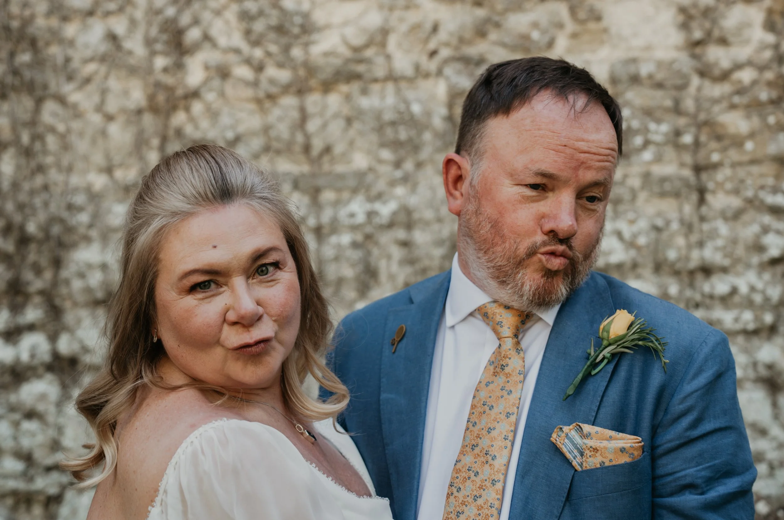 A smiling woman with blonde hair in a white dress standing next to a man with a beard in a blue suit, both dressed for a special occasion, with a stone wall in the background.