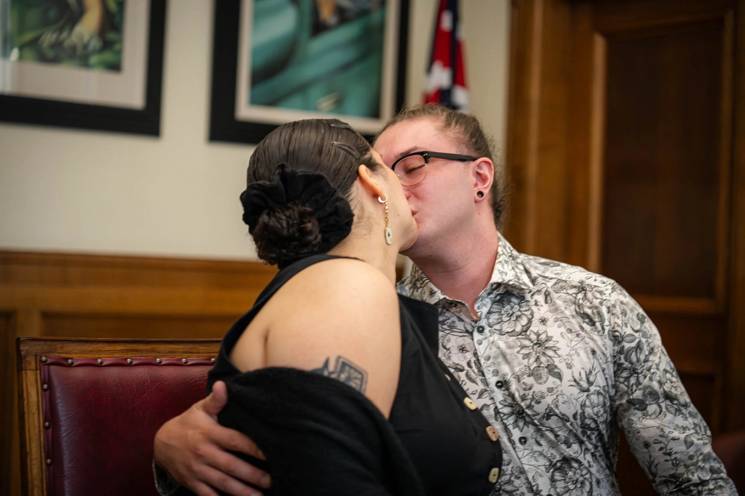 A couple sharing a kiss in a cozy indoor setting, with artworks on the wall behind them and a wood-paneled wall to the side.