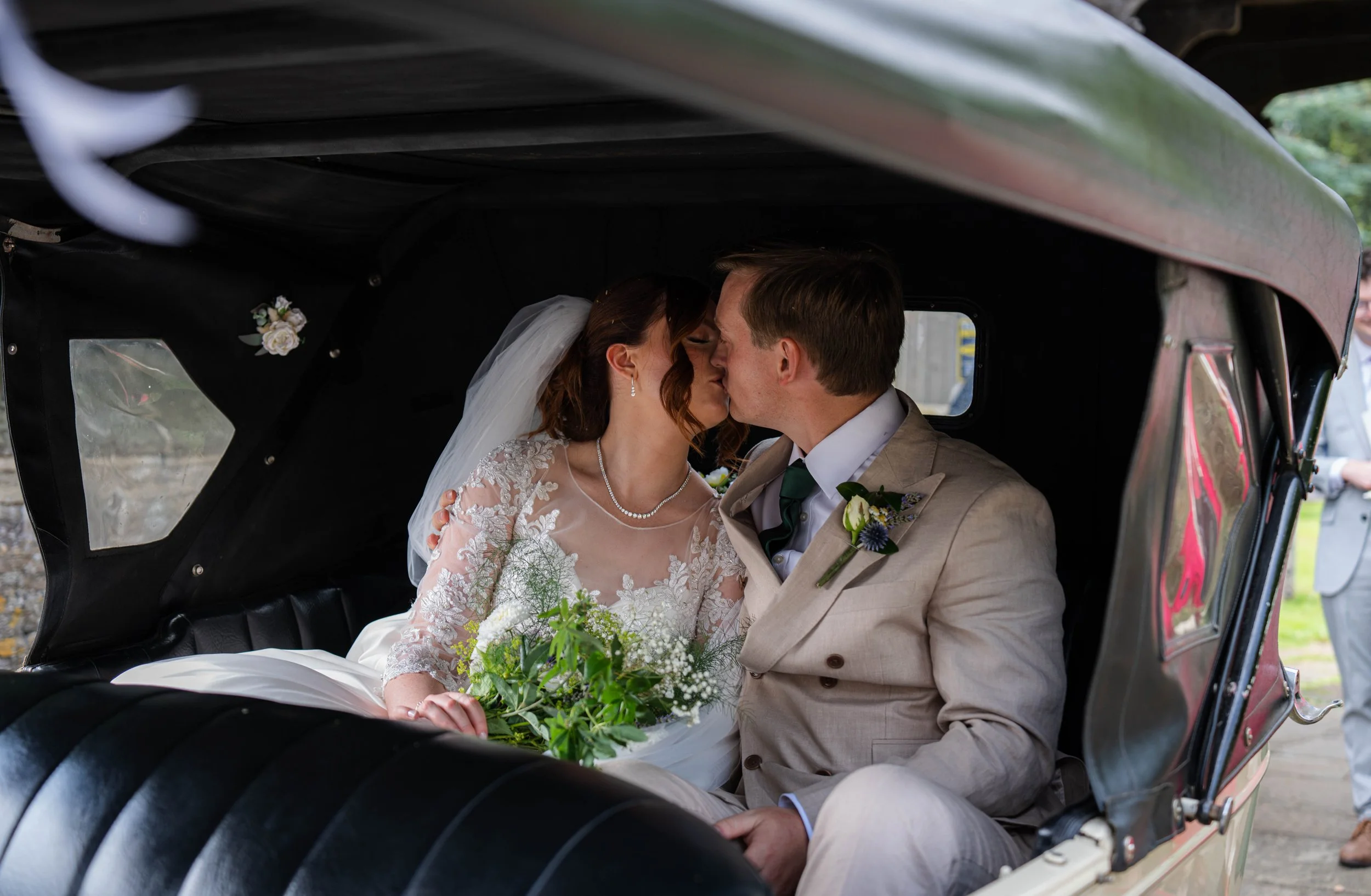 A bride and groom sharing a kiss inside a vehicle, with the bride holding a bouquet of flowers, during a wedding celebration.