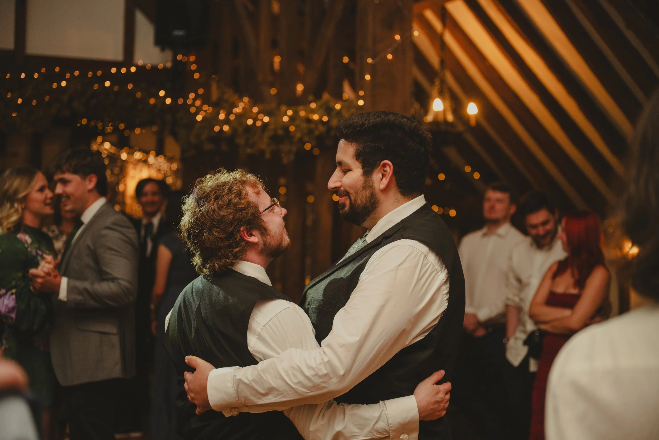 Two men are dancing closely with their arms around each other at a wedding reception, surrounded by other guests in a dimly lit venue decorated with warm string lights and candles.
