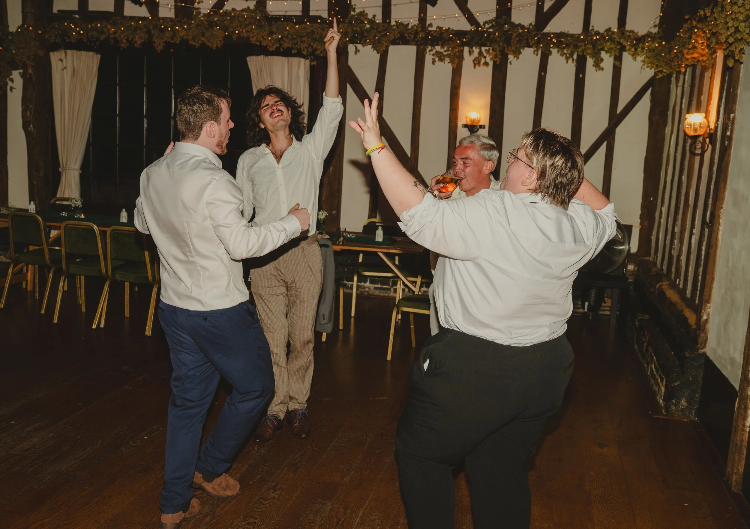 Four people dancing and having fun in a rustic indoor setting with wood beams and string lights, one wearing a white shirt with an arm raised, another with a drink, and others smiling and enjoying the moment.