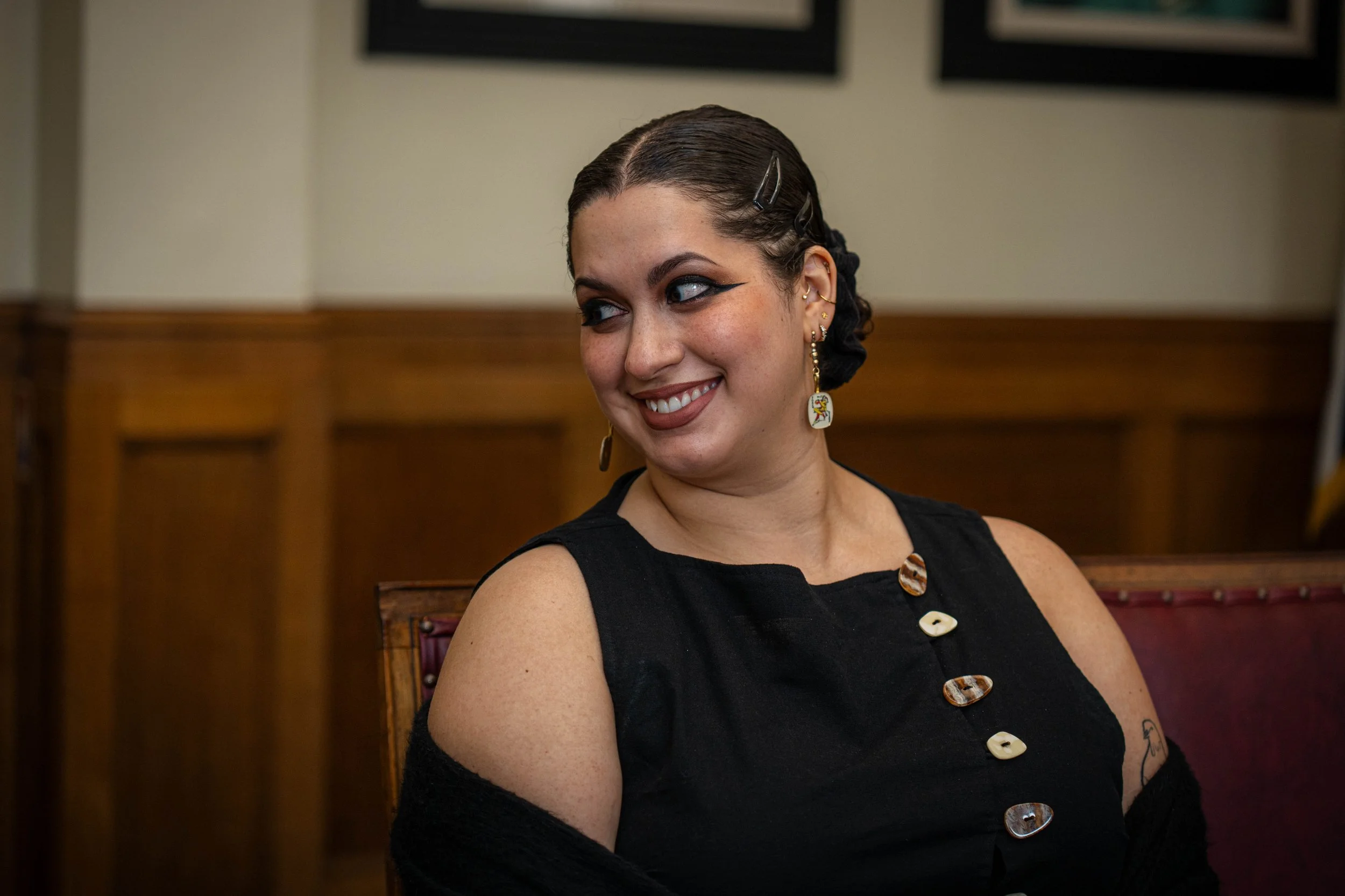A woman with dark hair styled with clips, smiling, wearing a black sleeveless top with decorative buttons, sitting on a wooden bench, with earrings and tattoos visible.