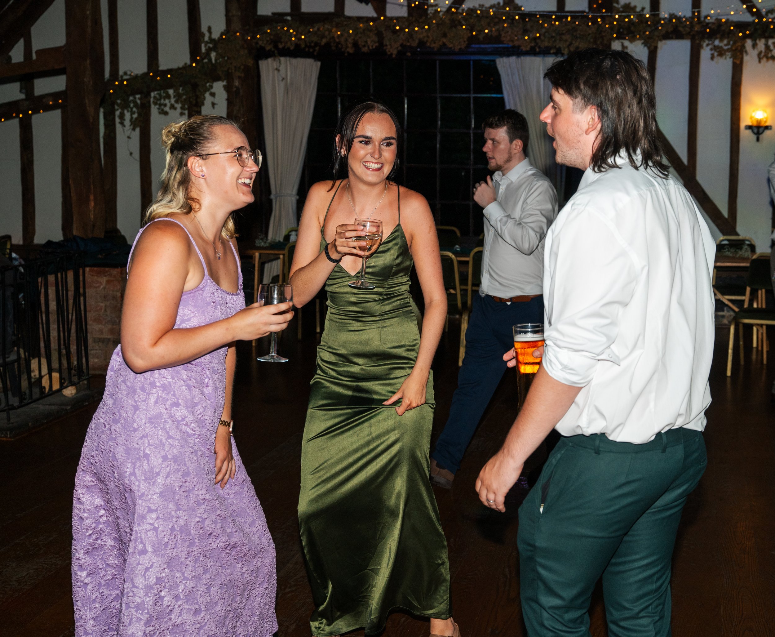 Group of four people, three women and one man, dressed up and socializing at a party or celebration in a rustic venue with string lights and curtains, smiling and holding drinks.