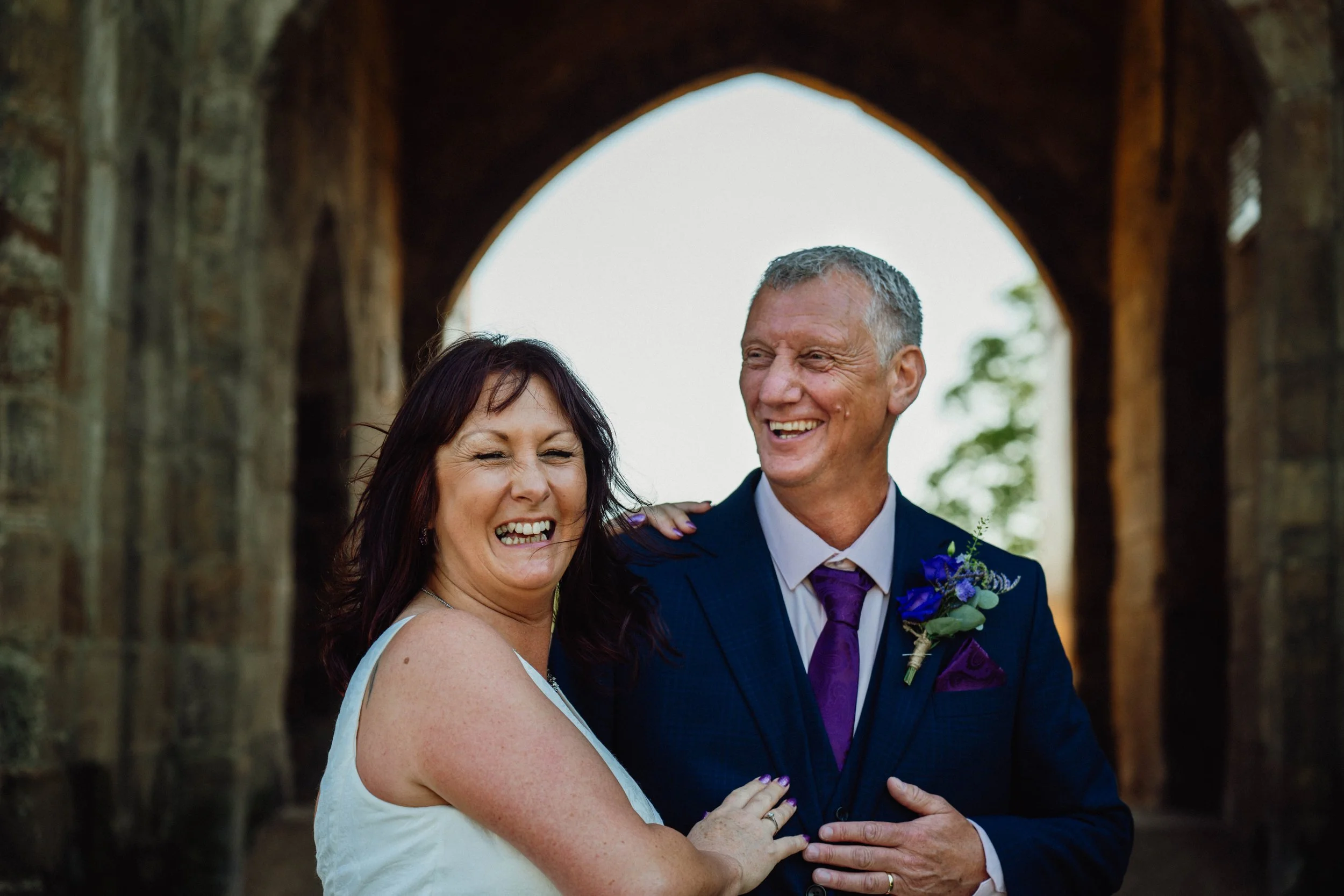 A smiling woman and man, dressed formally, standing close together outdoors with a stone archway in the background.