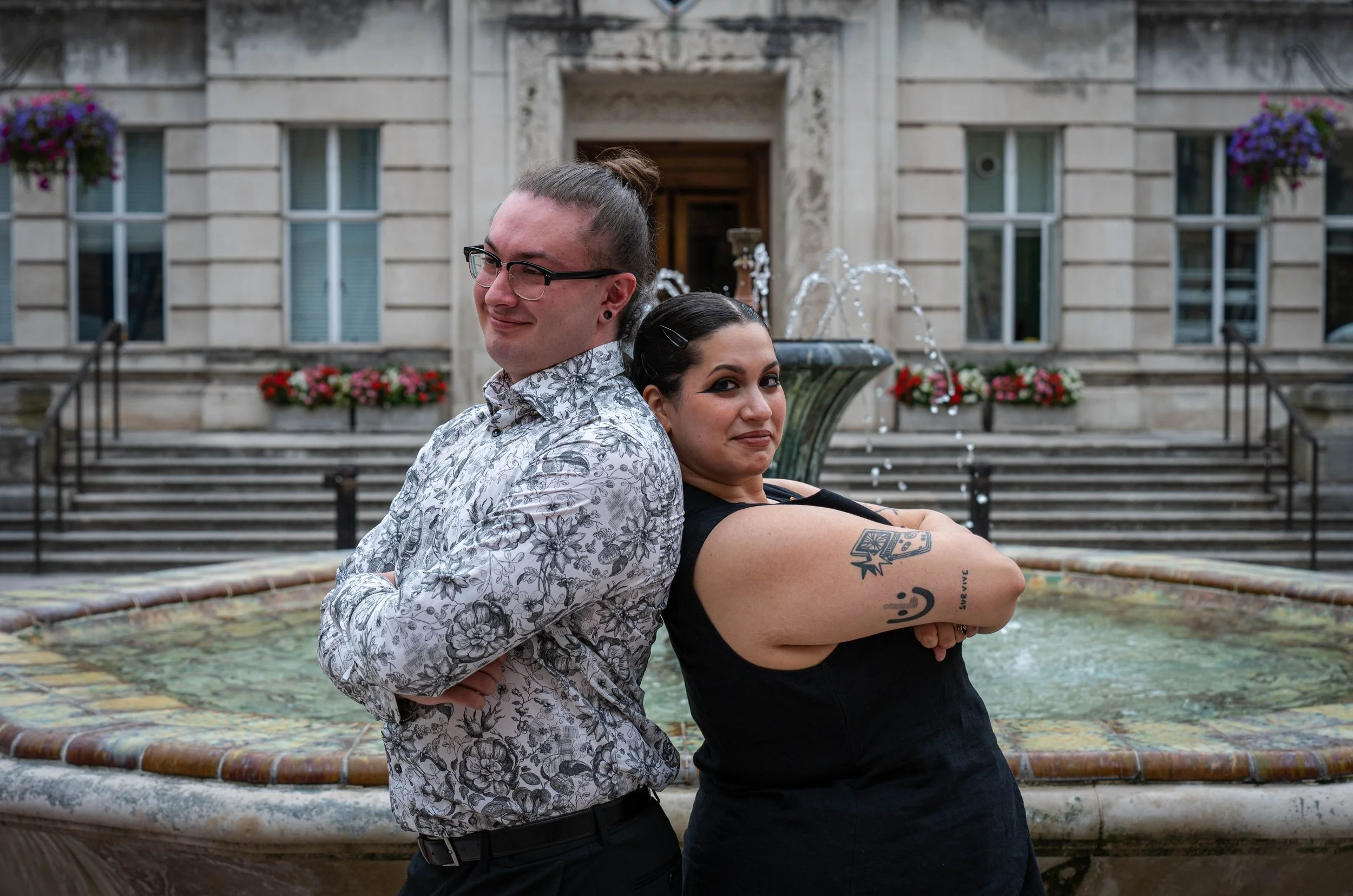 Two people, a man and a woman, stand with their backs pressed together in front of a fountain outdoors, with a building decorated with flowers in the background.