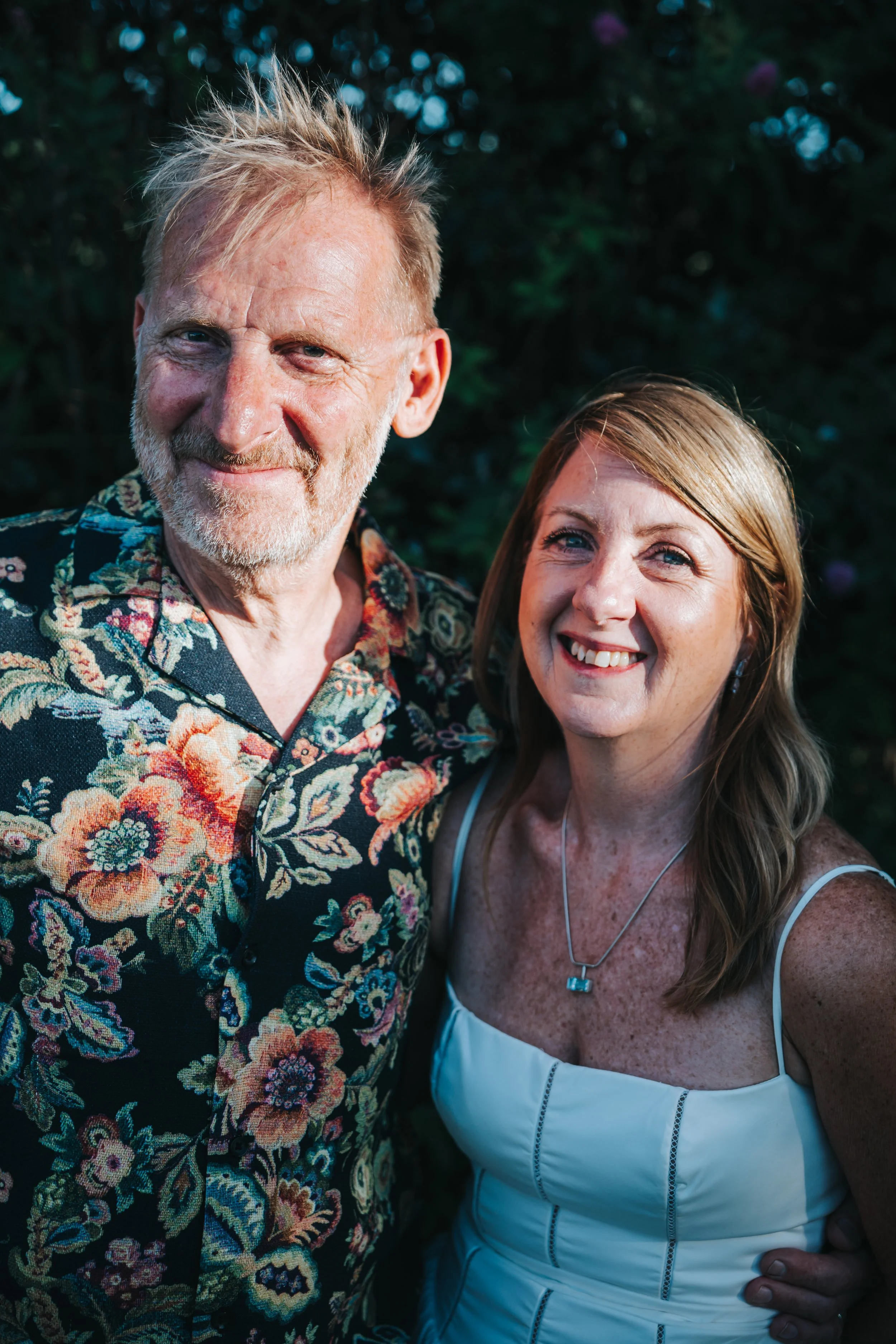 A man with a beard and mustache, wearing a floral shirt, standing next to a woman with long light brown hair, wearing a white dress with thin straps and a necklace, both smiling outdoors with greenery in the background.