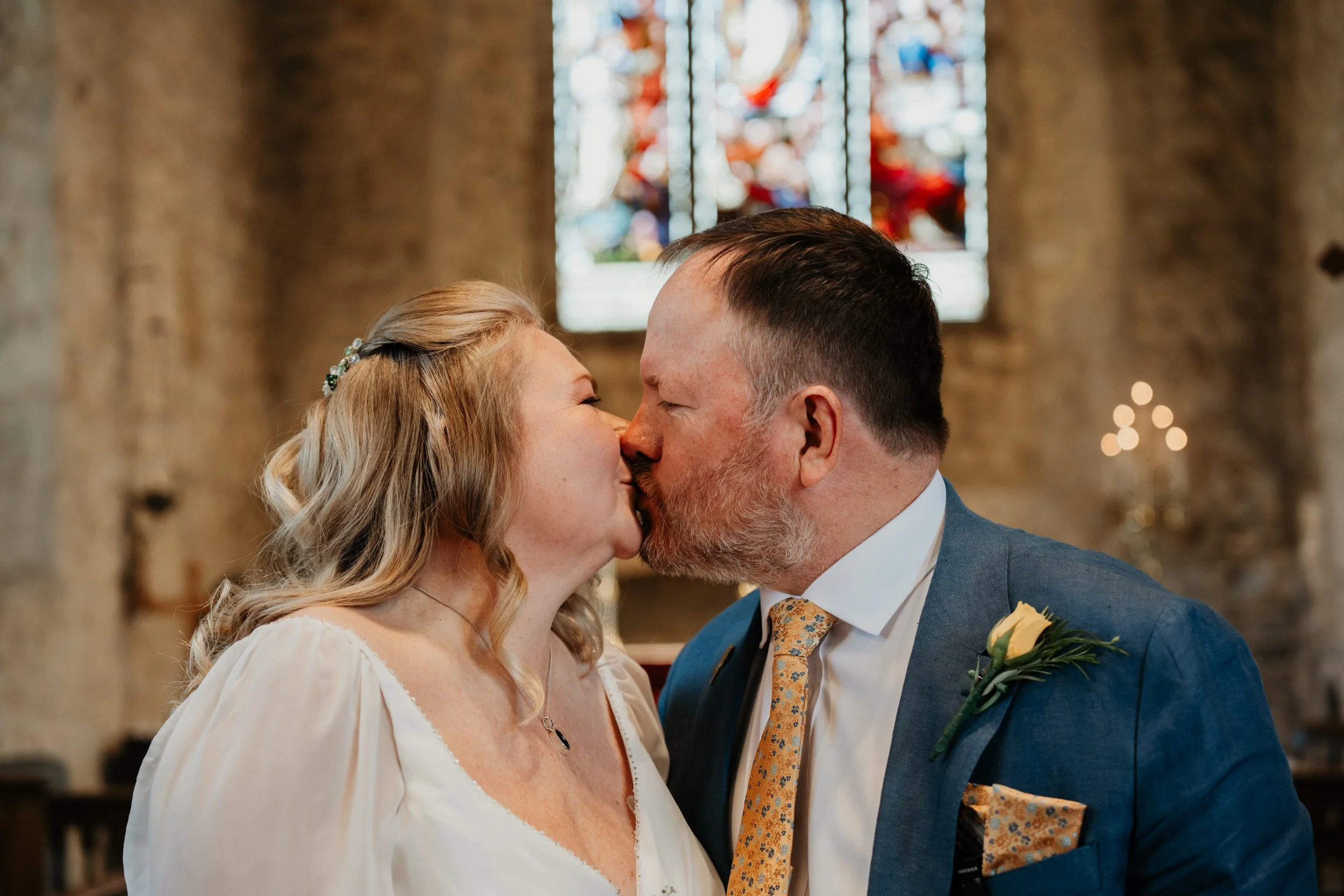 A bride and groom sharing a kiss during their wedding ceremony inside a church with stained glass windows in the background.
