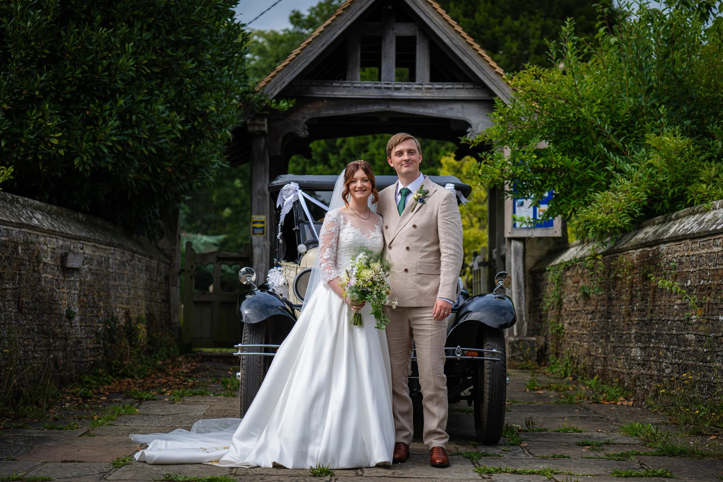 A bride and groom standing together in front of a vintage car decorated for a wedding, surrounded by greenery and an old wooden gate.