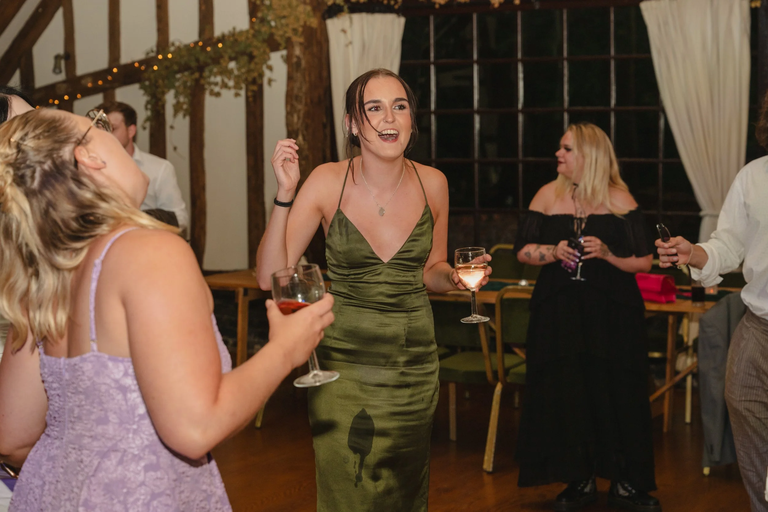 A woman with dark hair and a green dress laughing and holding a glass of white wine at a social gathering, surrounded by other women in a warmly lit room with wooden beams and windows.