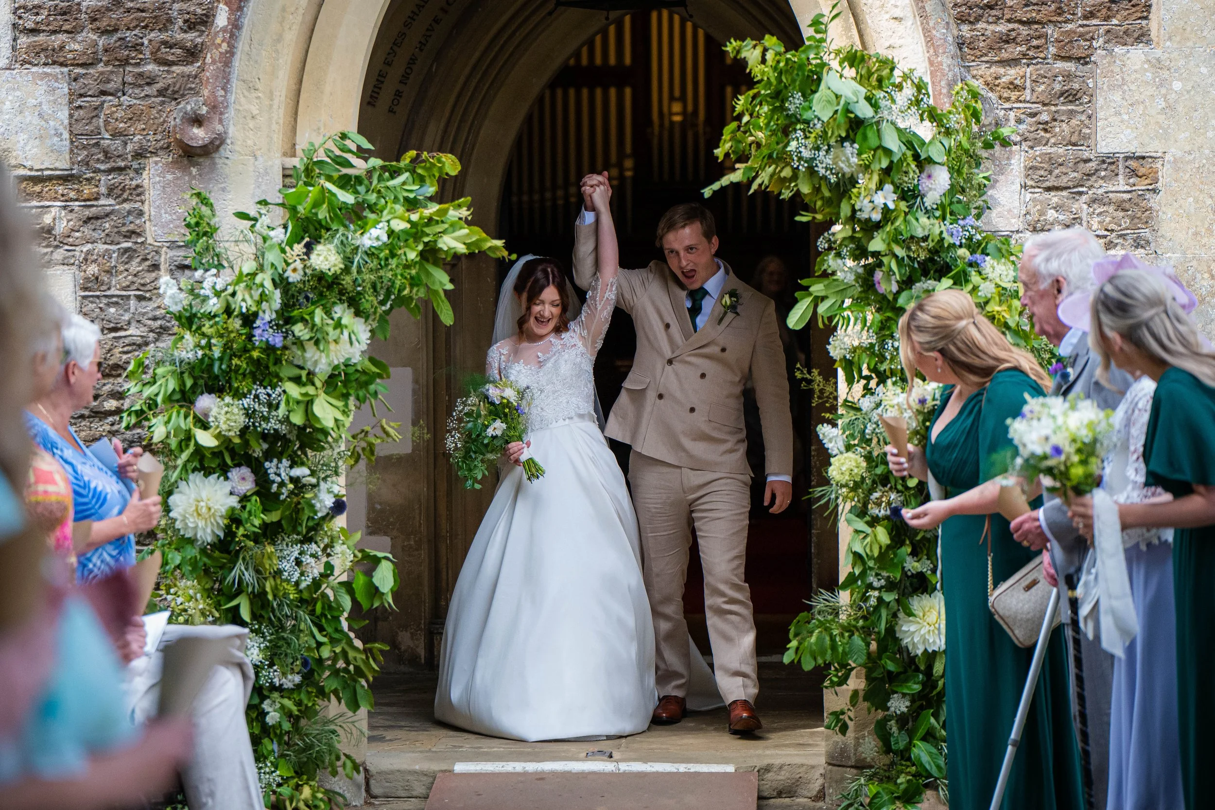 A newly married couple celebrating as they exit a church, surrounded by friends and family. The bride is wearing a white wedding dress and veil, holding a bouquet, and the groom is dressed in a tan suit with a boutonniere. They are holding hands and 