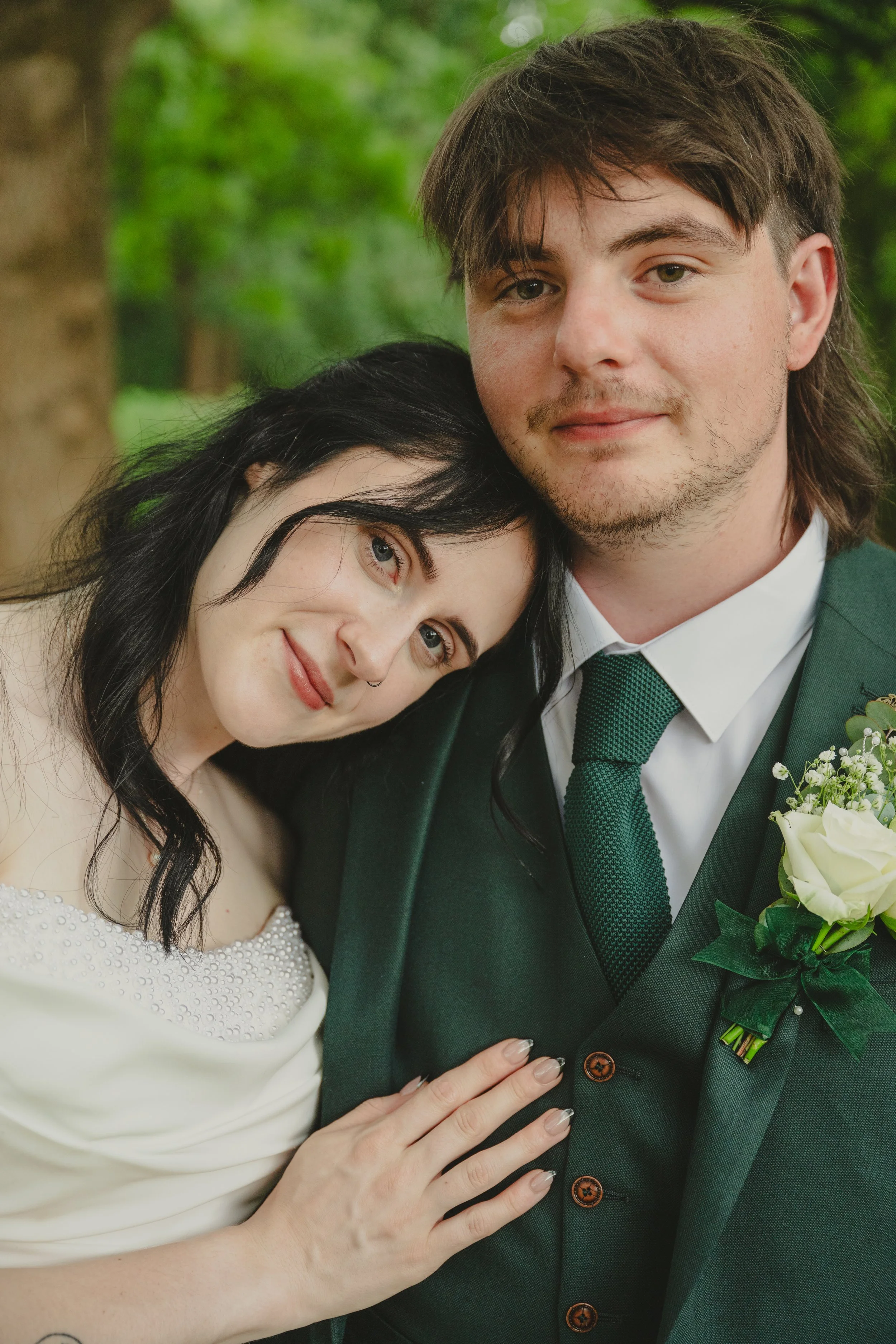 Close-up of young couple at a wedding, with woman resting her head on man's shoulder, both smiling, outdoors with green trees.