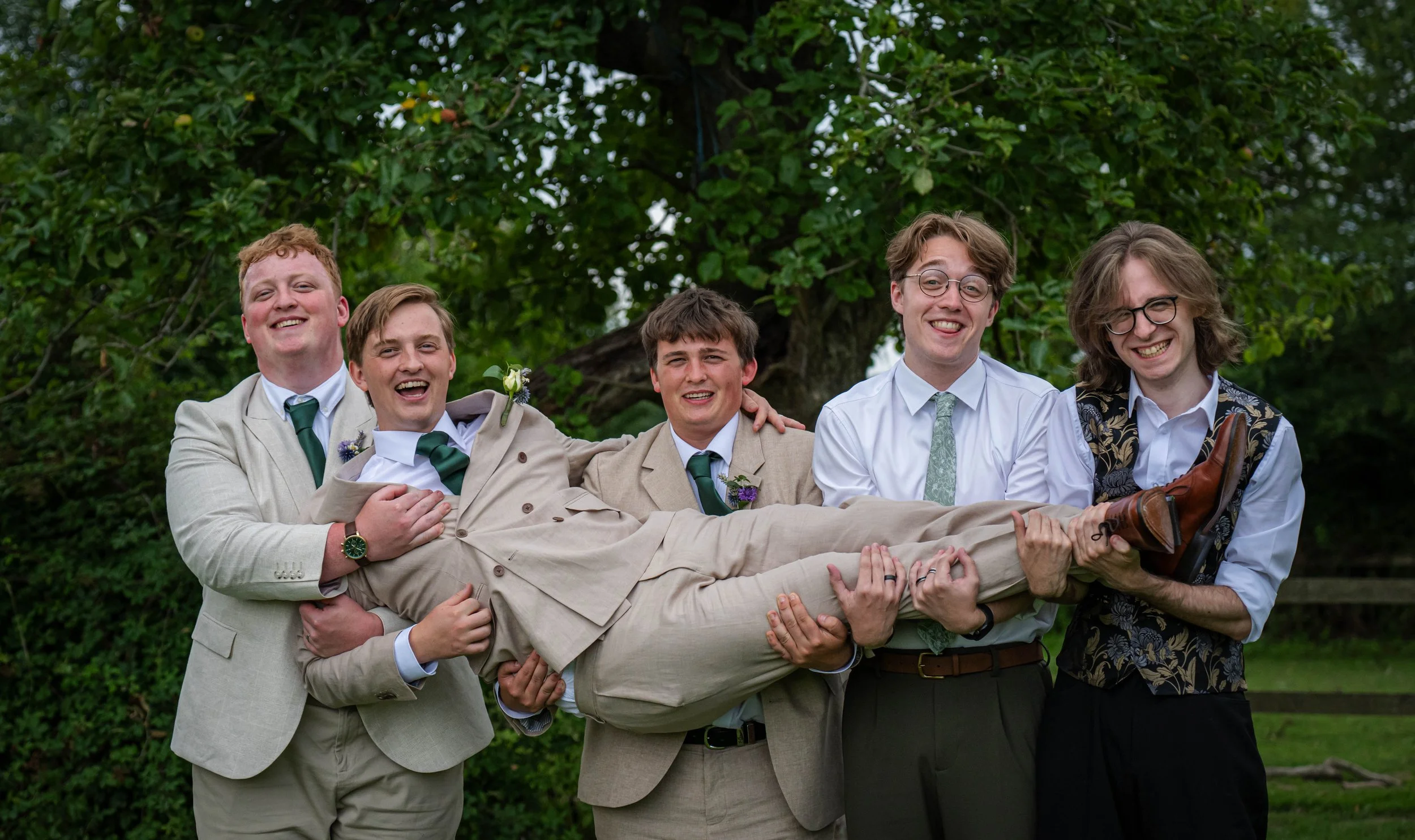 Group of five young men in suits lifting a person horizontally outdoors with greenery in the background.