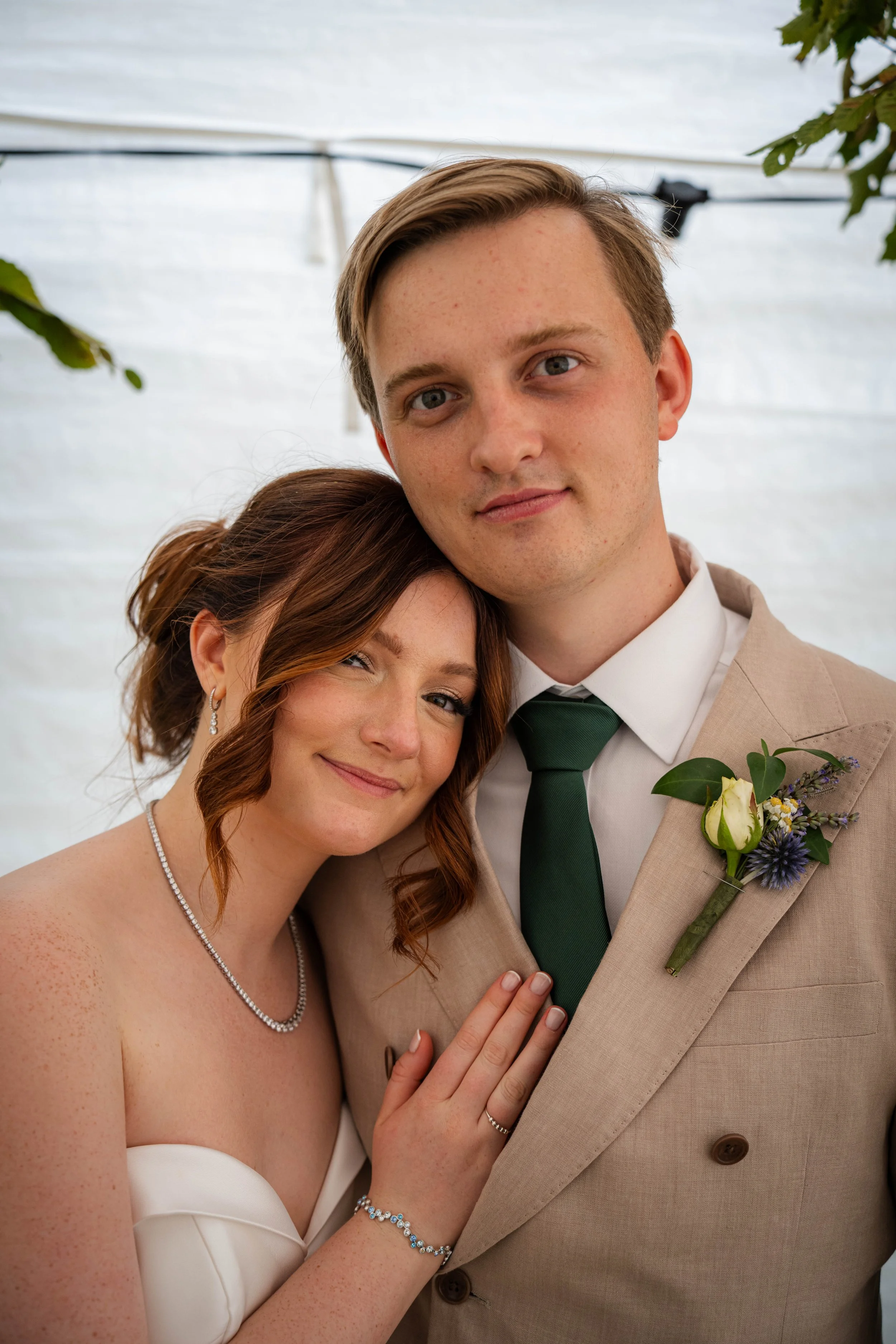 A bride and groom smiling, close-up, standing inside a white tent with greenery, the bride has red hair and wears a white strapless dress, a necklace, and bracelet, the groom has light brown hair, wears a beige suit with a dark green tie, and a bouto