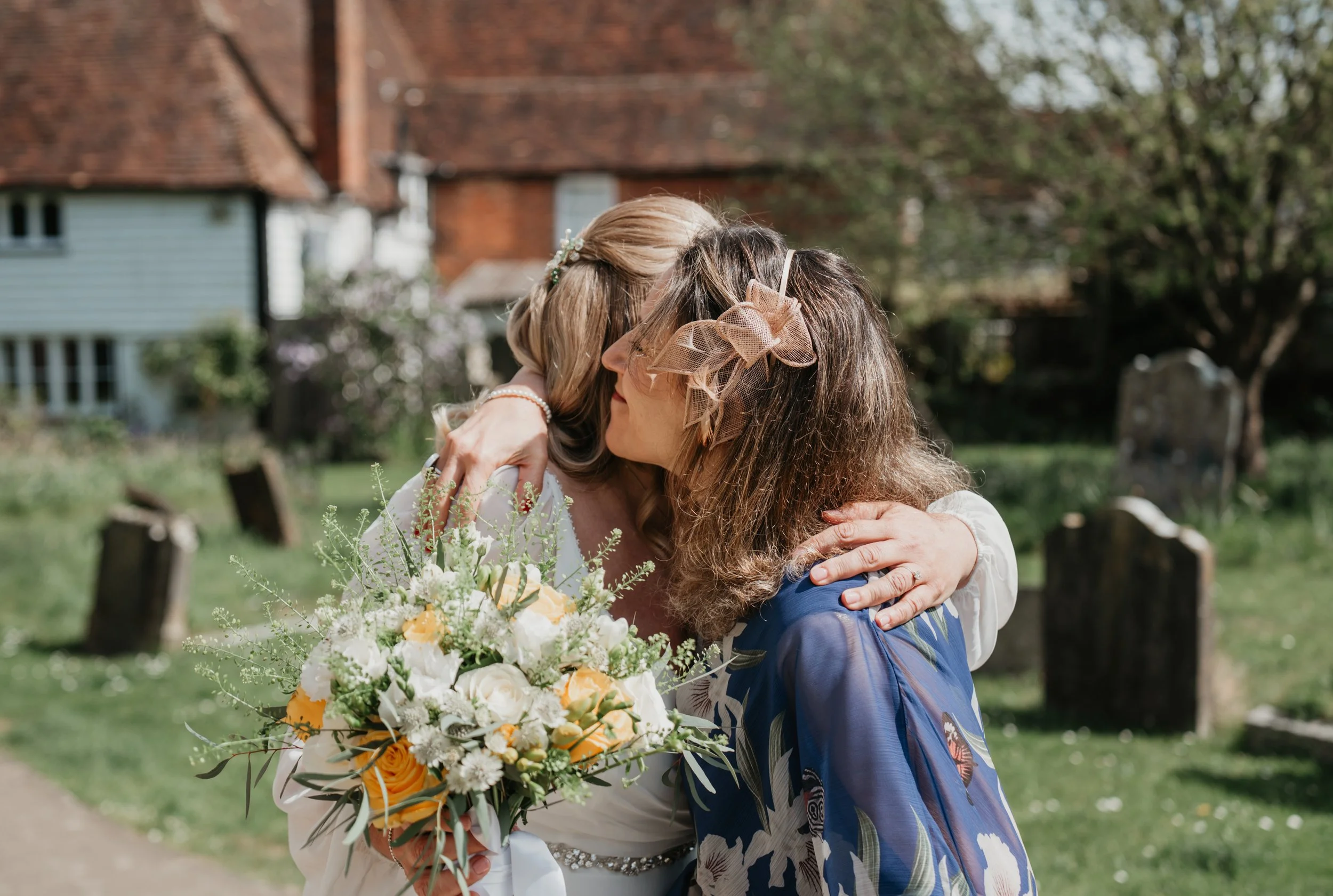 Two women hugging, one holding a large bouquet of flowers, outside in a garden area with a tree and houses in the background.