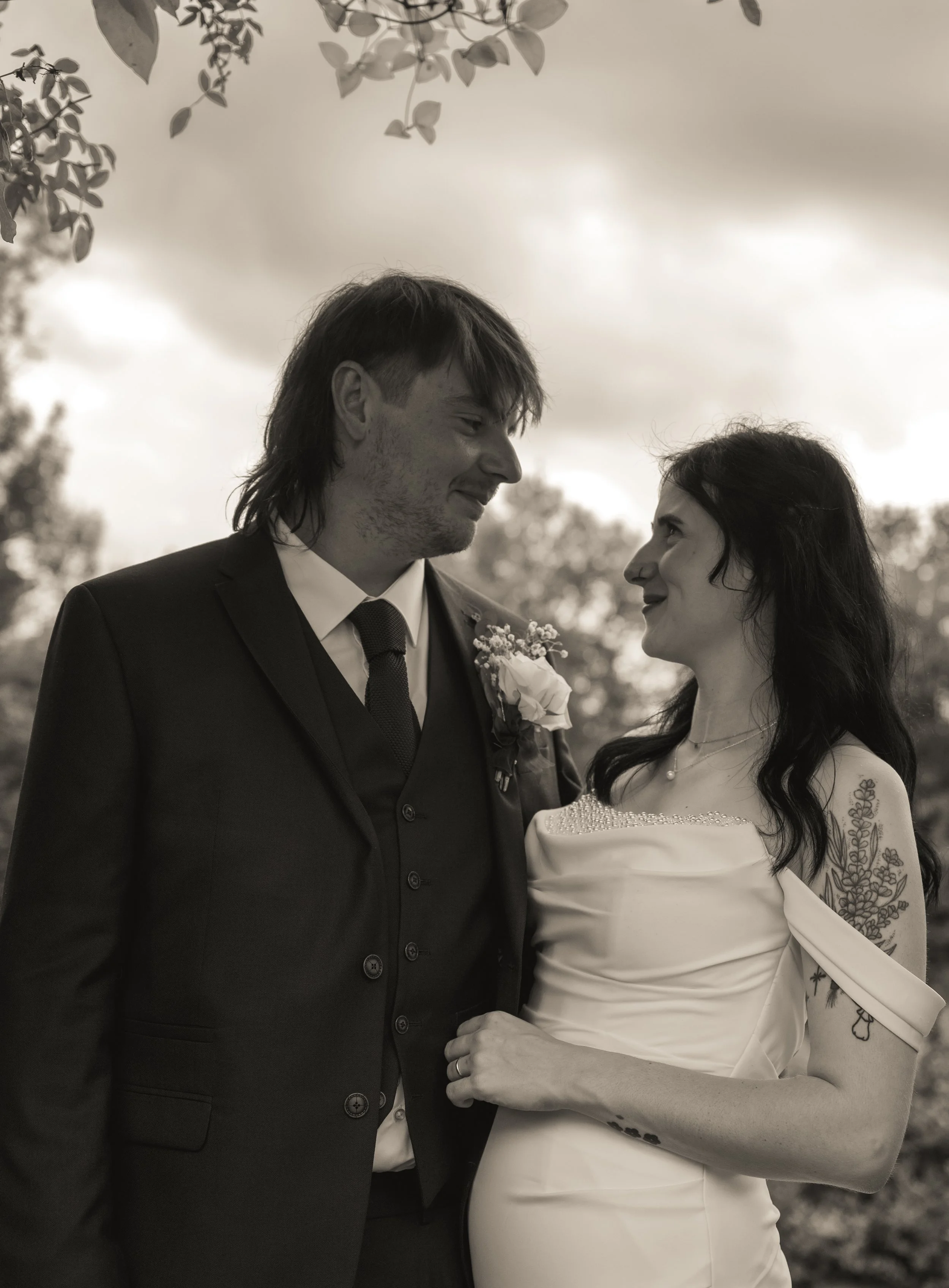 A black and white photo of a couple at their wedding. The man is wearing a suit with a boutonniere. The woman is wearing a wedding dress with lace details and a pearl necklace. They are facing each other outdoors, holding hands and looking into each 