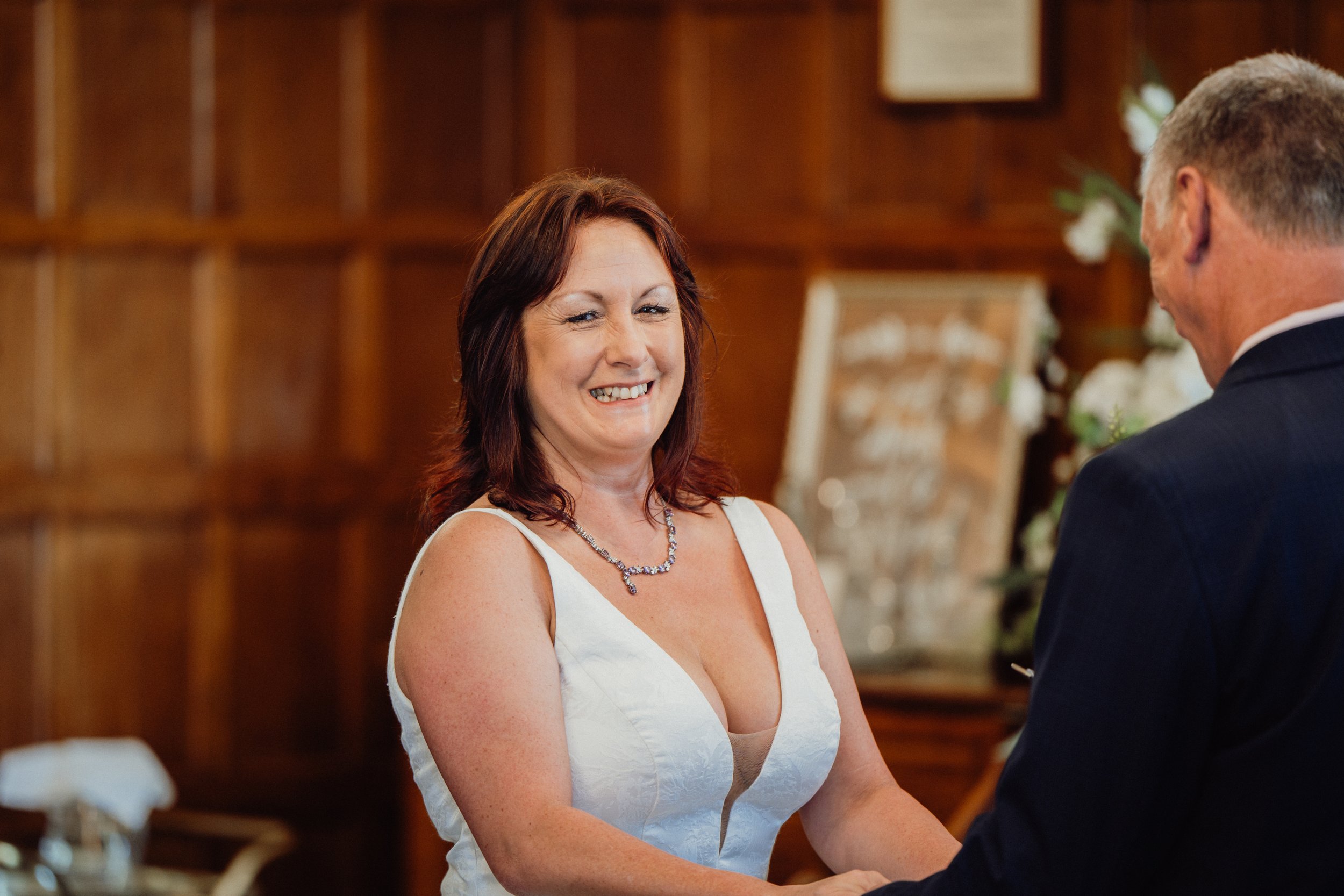 A woman in a white dress at a wedding ceremony, smiling while holding hands with a man in a dark suit. The background features wooden walls and blurred flowers.