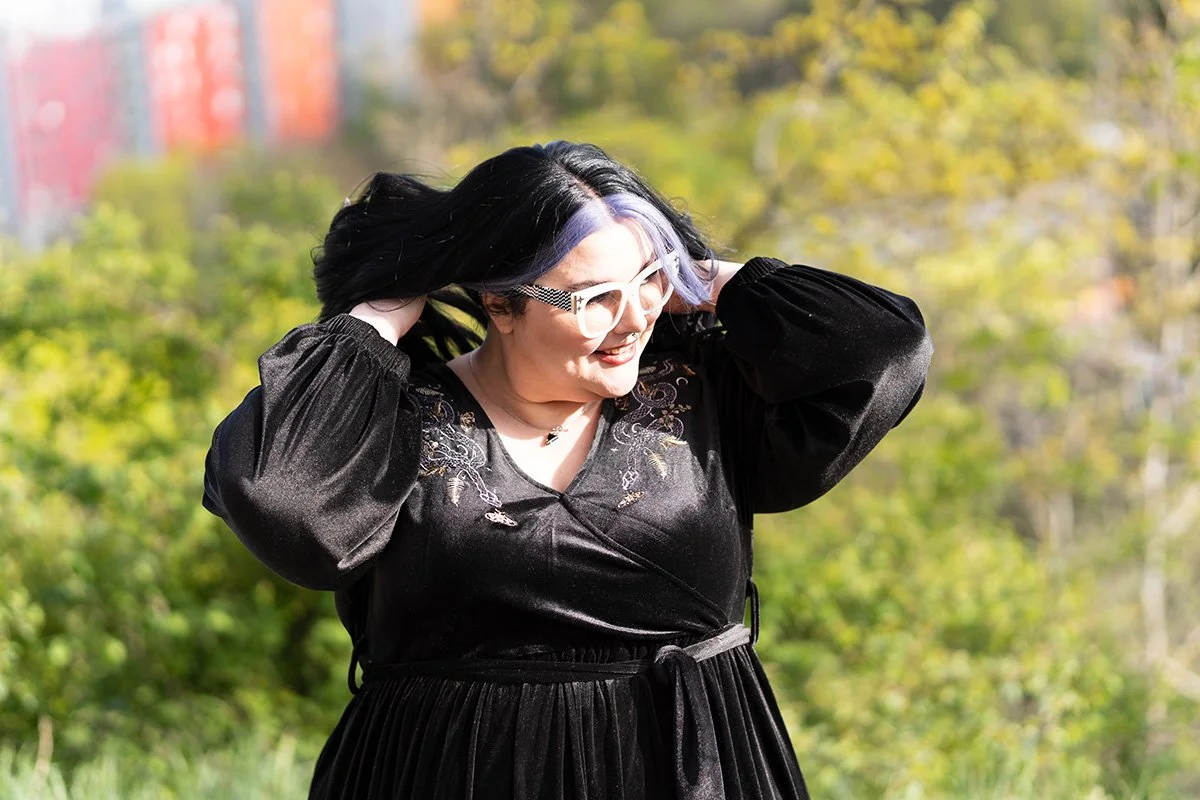 A woman with glasses and black hair with purple streaks smiling outdoors in a park with green trees in the background, wearing a black dress with embroidery.