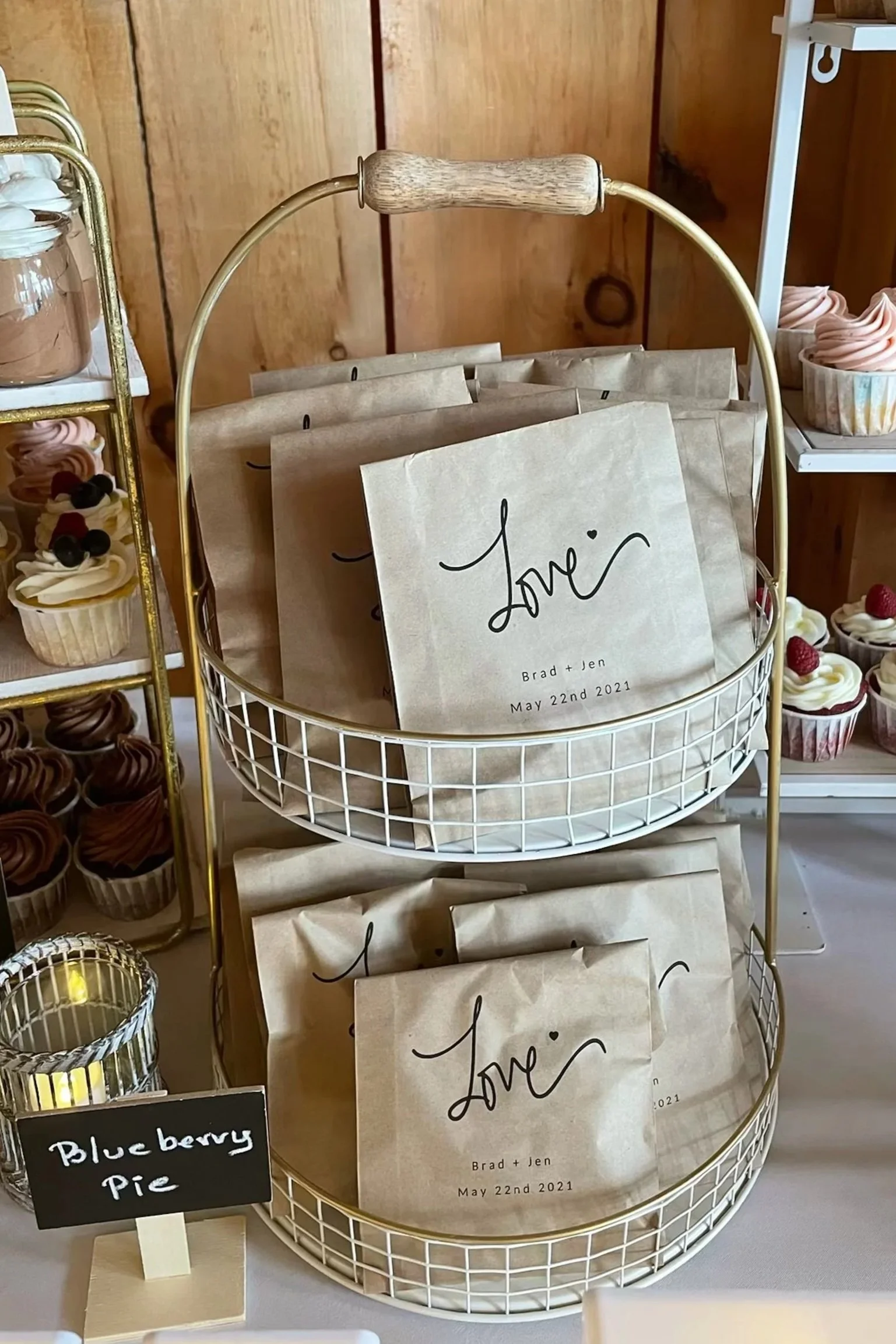 Wedding favor bags labeled 'Love' with the names Brad and Jen and the date May 22nd, 2021, displayed on a white wire basket at a dessert table with cupcakes and a small sign reading 'Blueberry Pie'.
