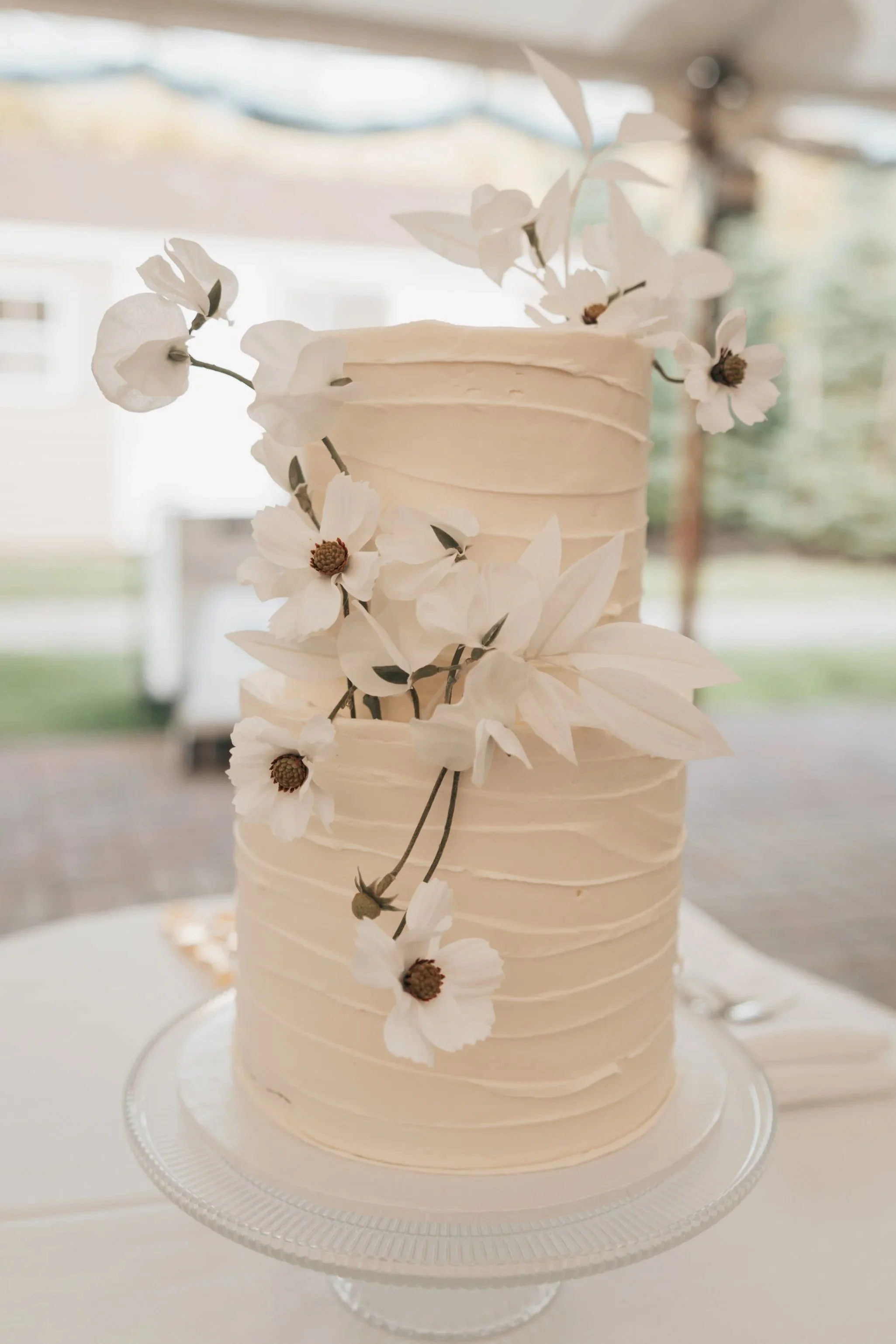 A white, multi-layered cake decorated with white flowers and foliage, displayed on a glass cake stand.