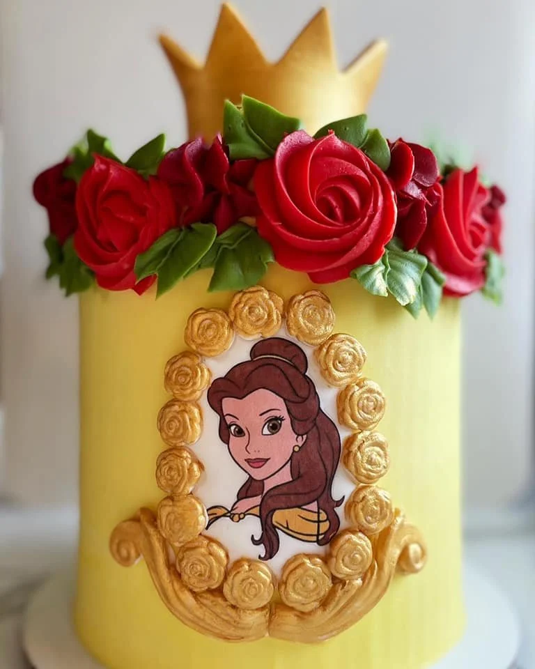 A yellow birthday cake decorated with red roses, green leaves, a gold crown, and an image of Belle from Disney's Beauty and the Beast, surrounded by gold roses.