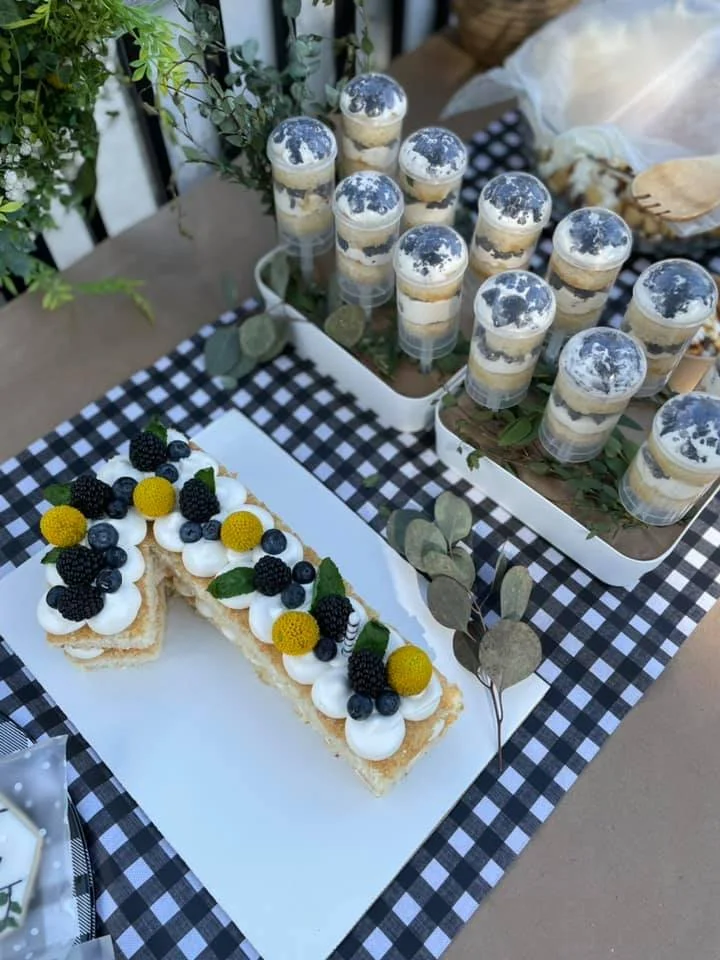 Dessert table with berry-topped pastry and layered parfaits topped with cookies, blackberries, raspberries, blueberries, and mint leaves, on a checkered tablecloth.