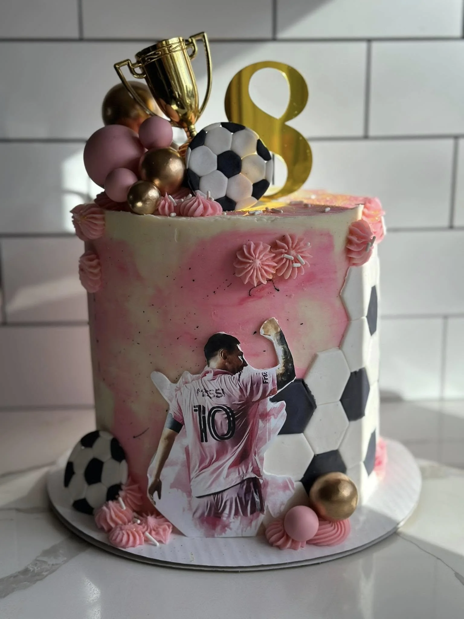 A decorated cake with pink and white icing, soccer-themed decorations including a paper cutout of a soccer player, a black and white soccer ball, and a gold trophy topper, along with pink, gold, and purple decorative balls, and a big gold number thre