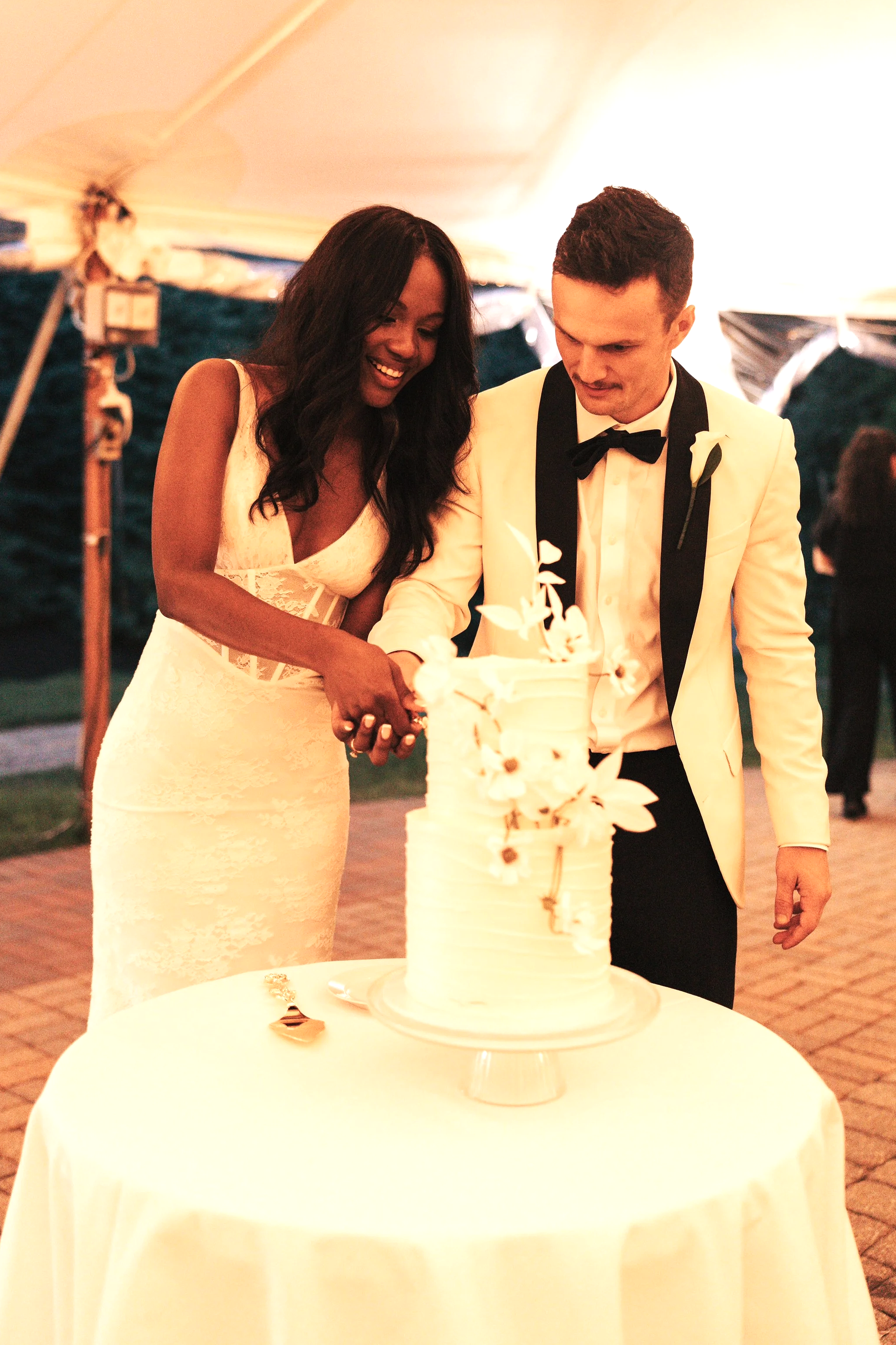 A bride and groom cutting a wedding cake at their reception in a tent, with warm lighting and a white tablecloth on the table.
