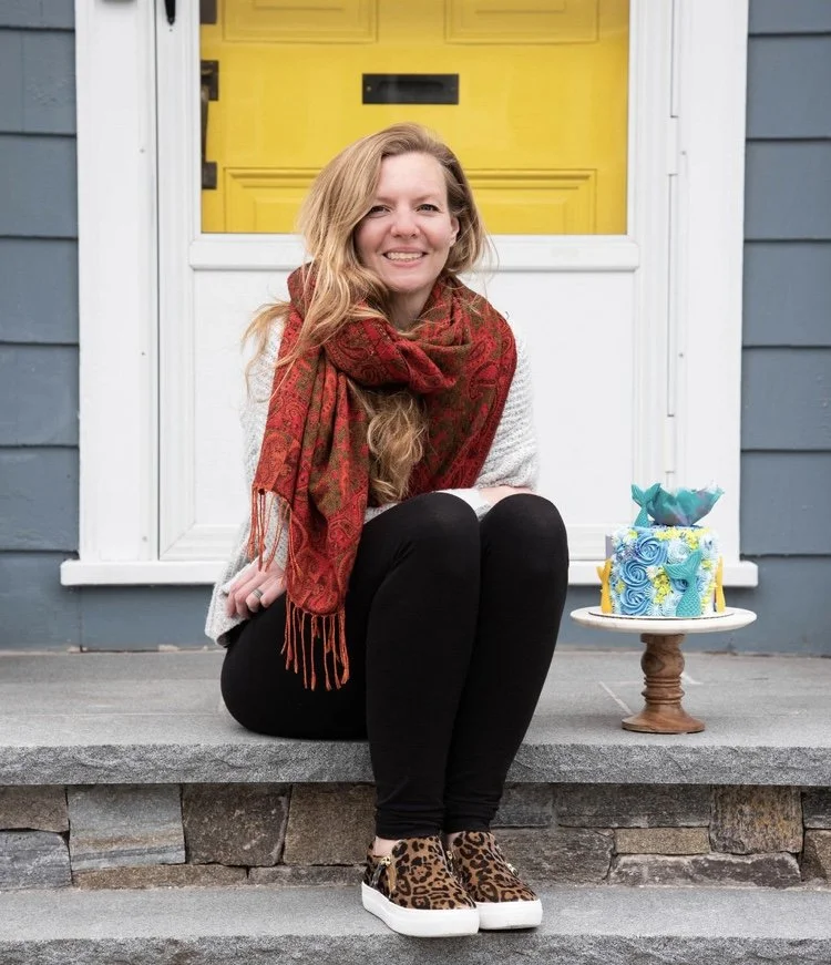 A woman sitting on a front porch step with a decorated cake on a stand next to her, in front of a house with a yellow door and gray siding.