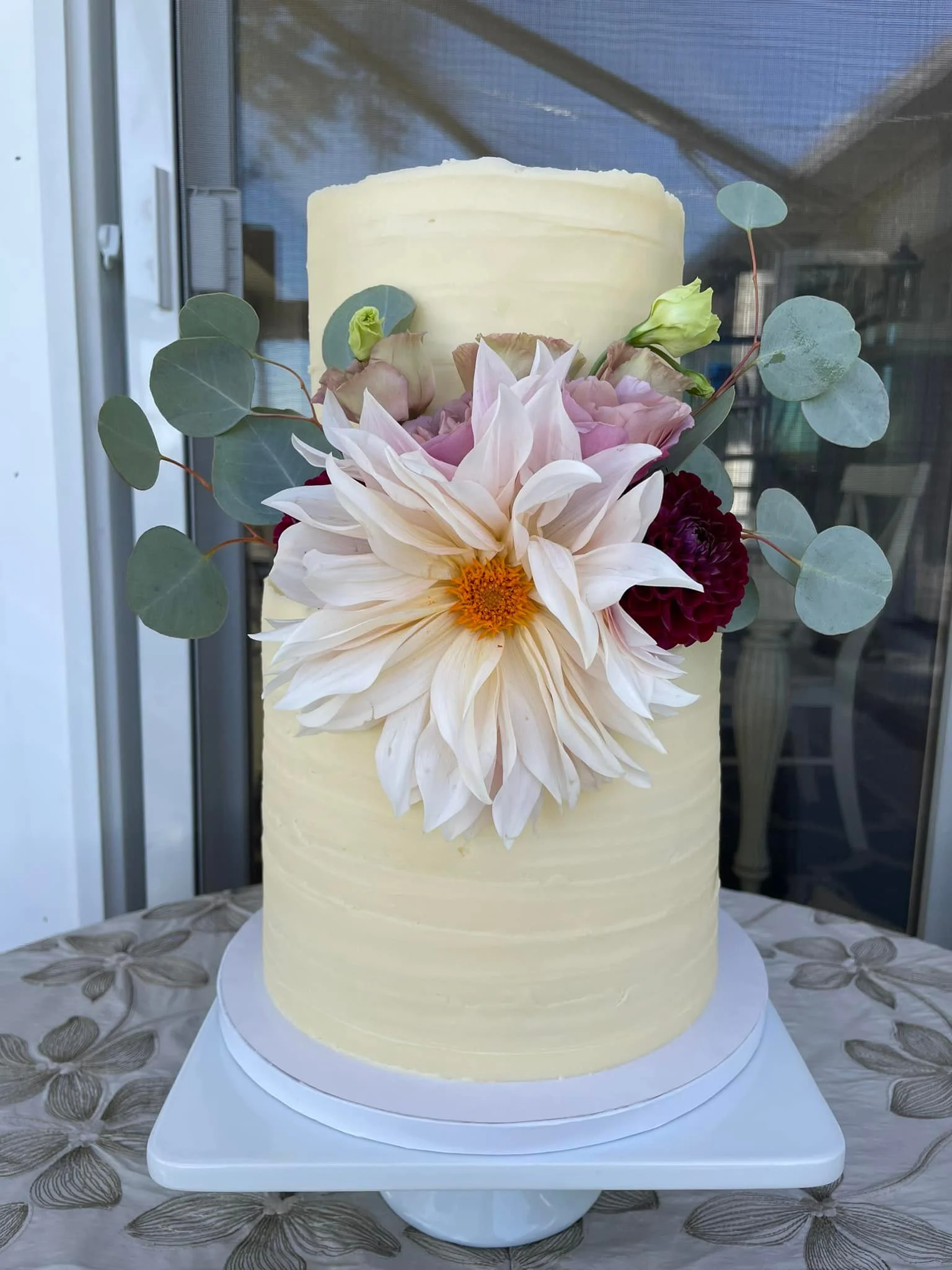 Cream-colored, tall cylindrical wedding cake decorated with fresh flowers including a white dahlia, pink roses, dark red flowers, eucalyptus leaves, and small light green buds, placed on a white cake stand.