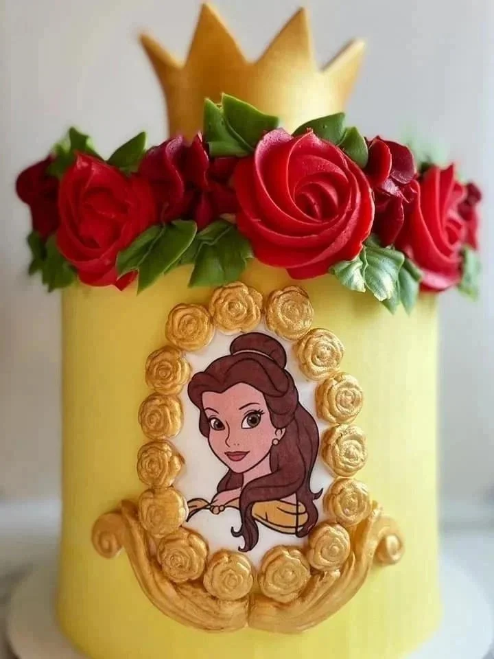A yellow birthday cake decorated with red roses, green leaves, a paper princess character resembling Belle from Beauty and the Beast, and a golden crown on top.