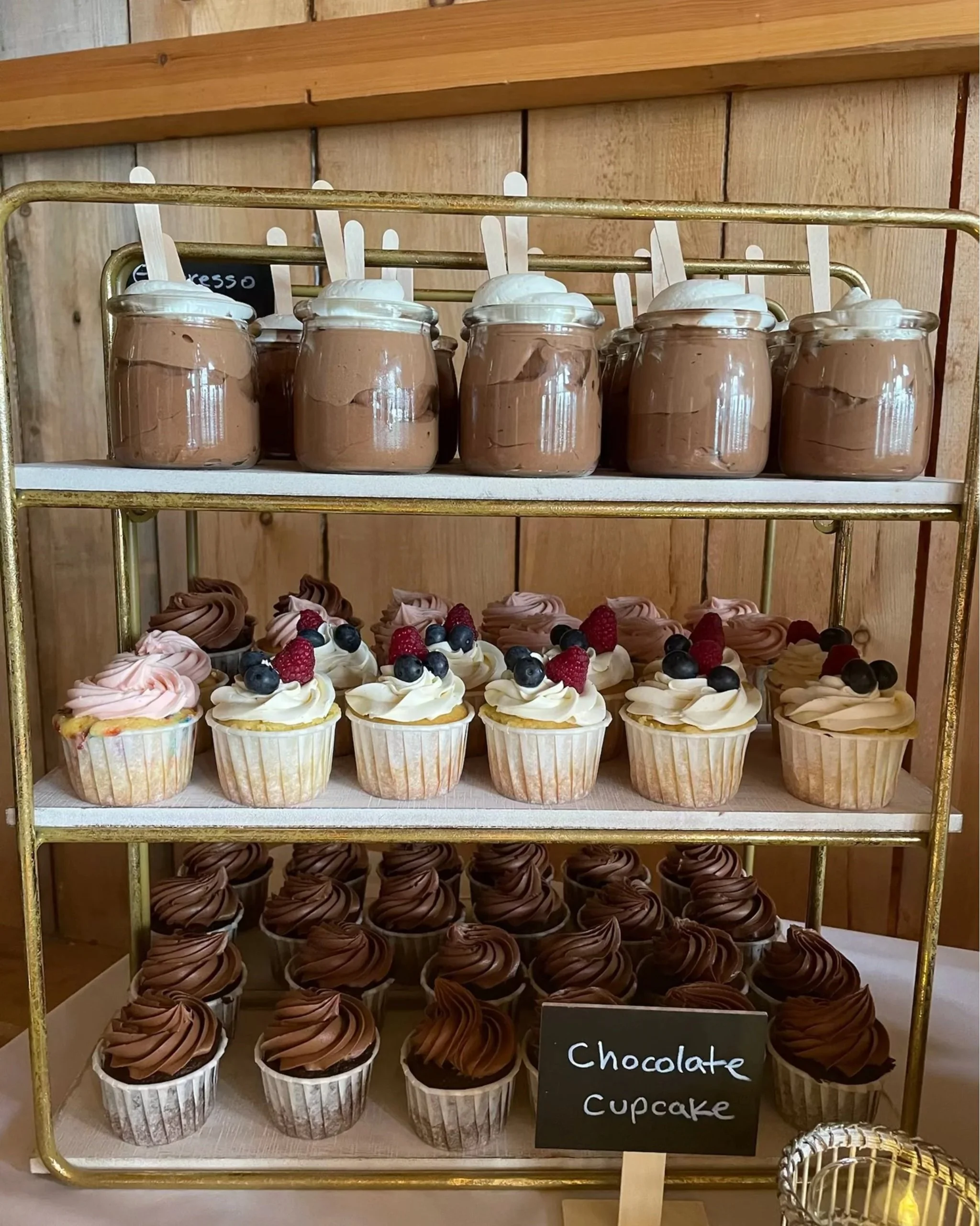 A three-tiered dessert display features jars of chocolate mousse on the top, decorated cupcakes with fruit and frosting in the middle, and chocolate cupcakes with swirled frosting on the bottom. A small chalkboard sign reads 'Chocolate Cupcake'.