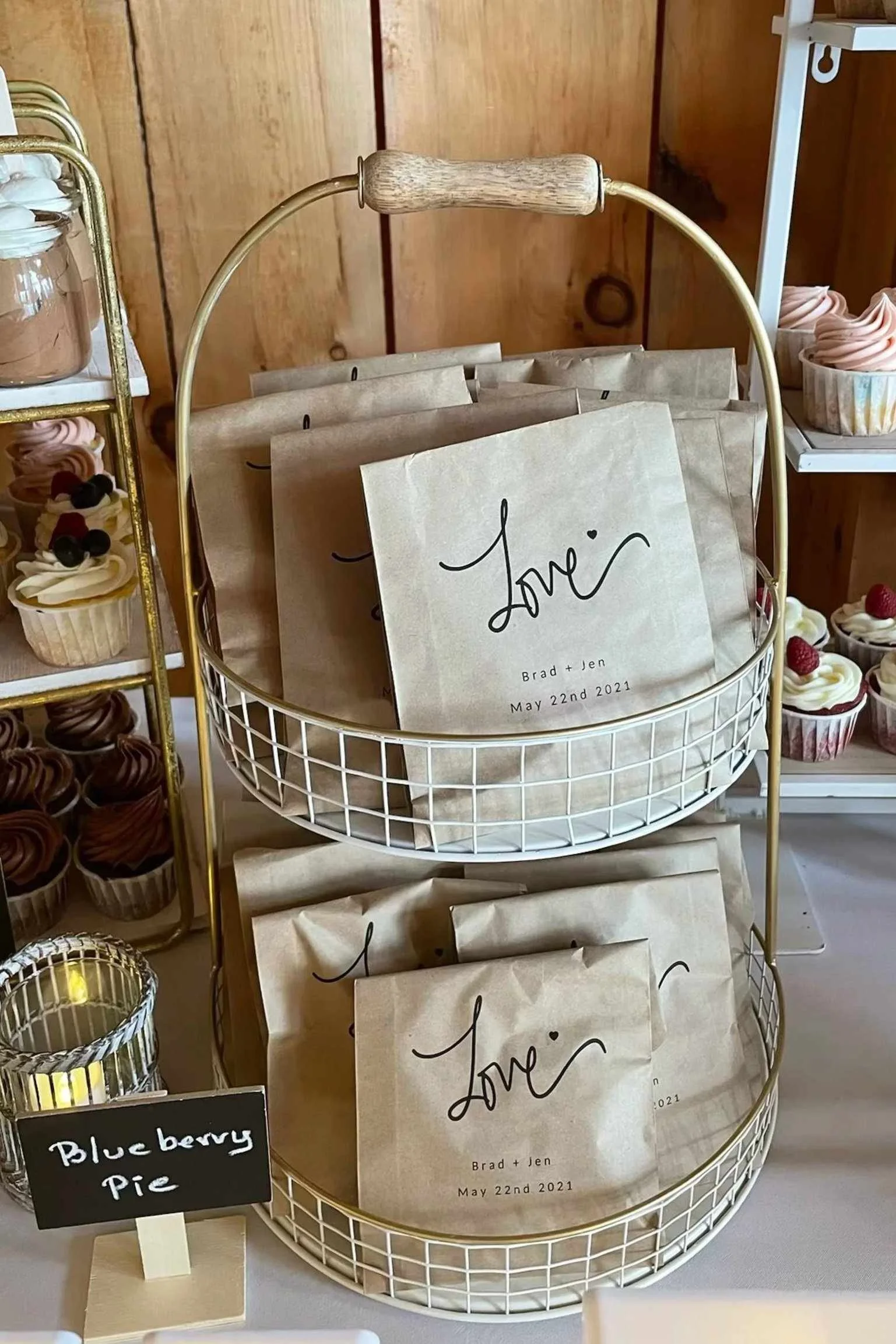 A white wire basket displays paper goodie bags, each with the word 'Love' written on them, along with names and date, against a wooden background. Shelves to the right contain cupcakes with pink frosting and fruit toppings. A small sign indicating blueberry pie is on the bottom left.