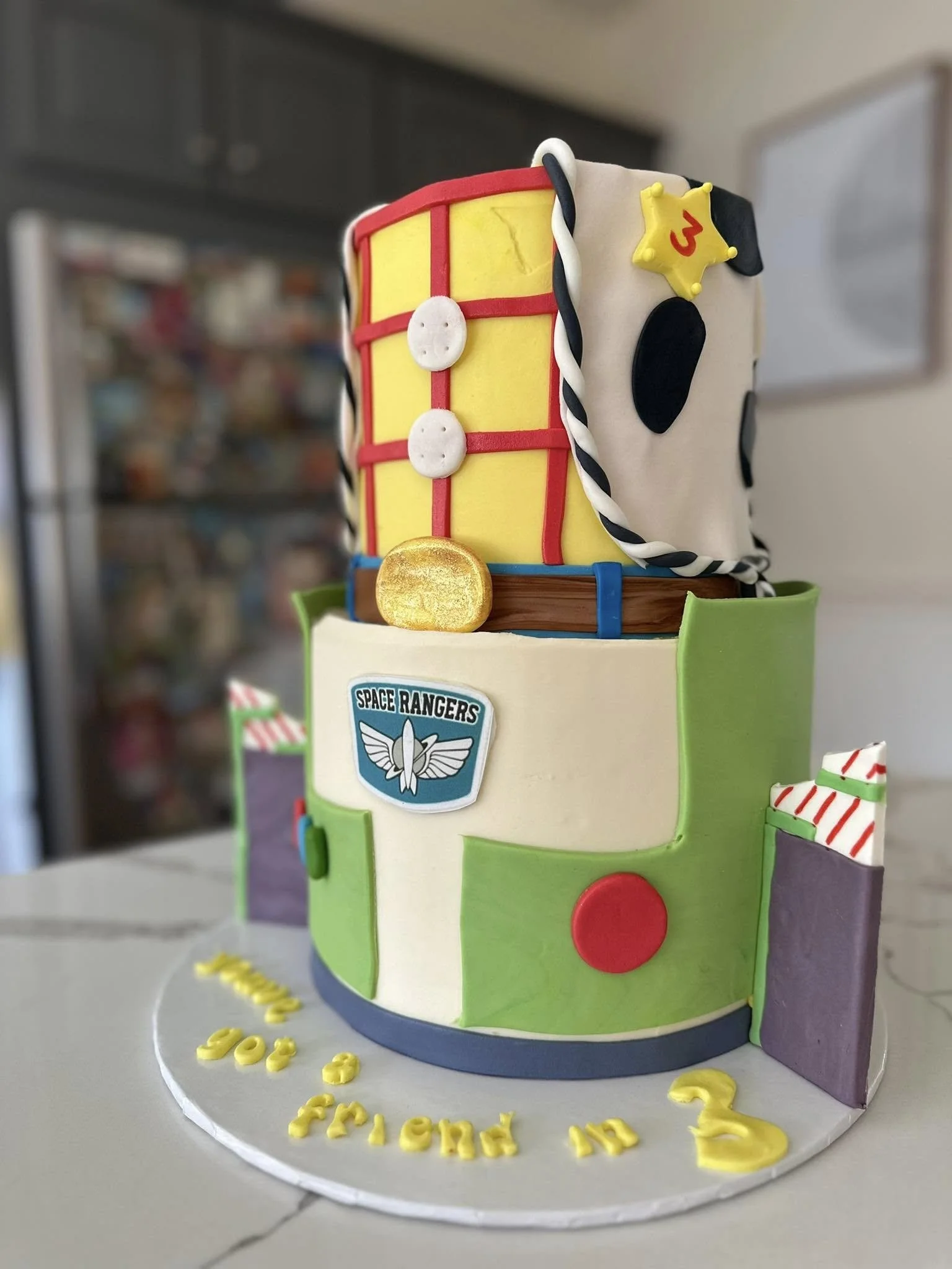A two-tiered cake decorated with a Toy Story theme, featuring Sheriff Woody's sheriff badge, a Woody hat with a star, and a Space Rangers logo, with colorful faux building blocks and a message that reads "Happy 3rd Friend in 3," celebrating a child's