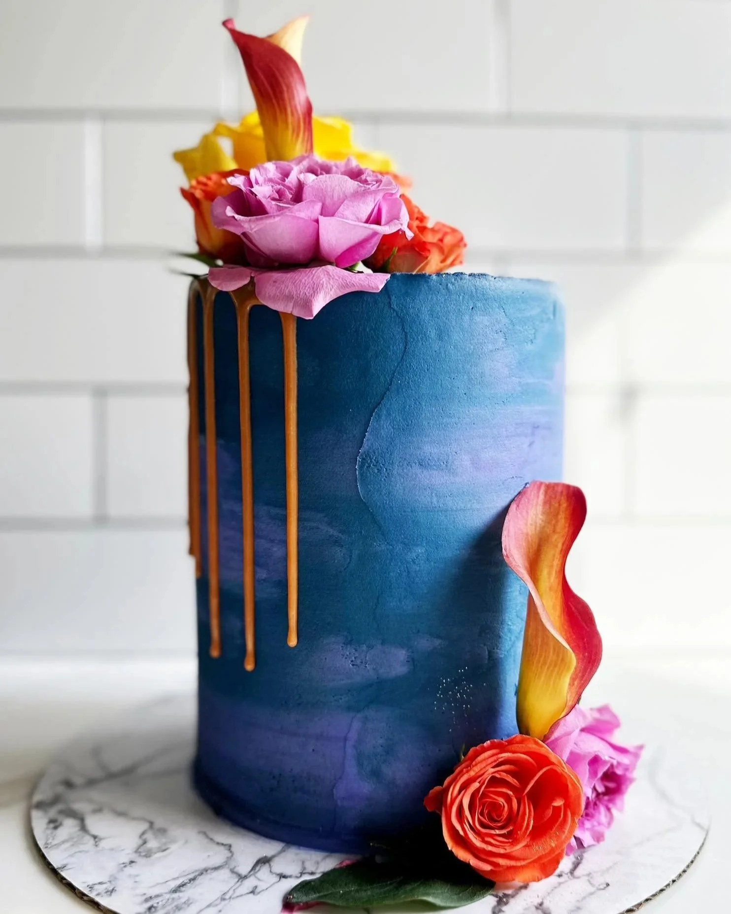 A tall blue cake decorated with fresh pink, orange, and yellow roses and petals, with gold drip accents on the side, placed on a marble-patterned cake board.