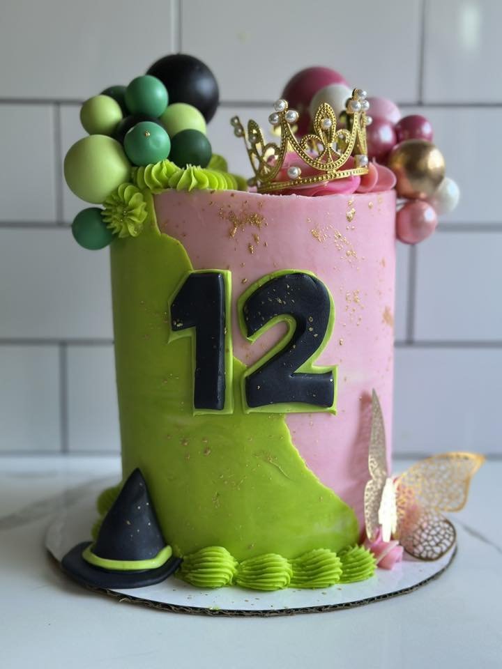 A colorful birthday cake decorated with a pink and green color scheme, adorned with a gold crown, edible pearls, and various spherical decorations. The cake displays a large number '12' on the front, with a small black witch hat at the base and a delicate butterfly on the side.