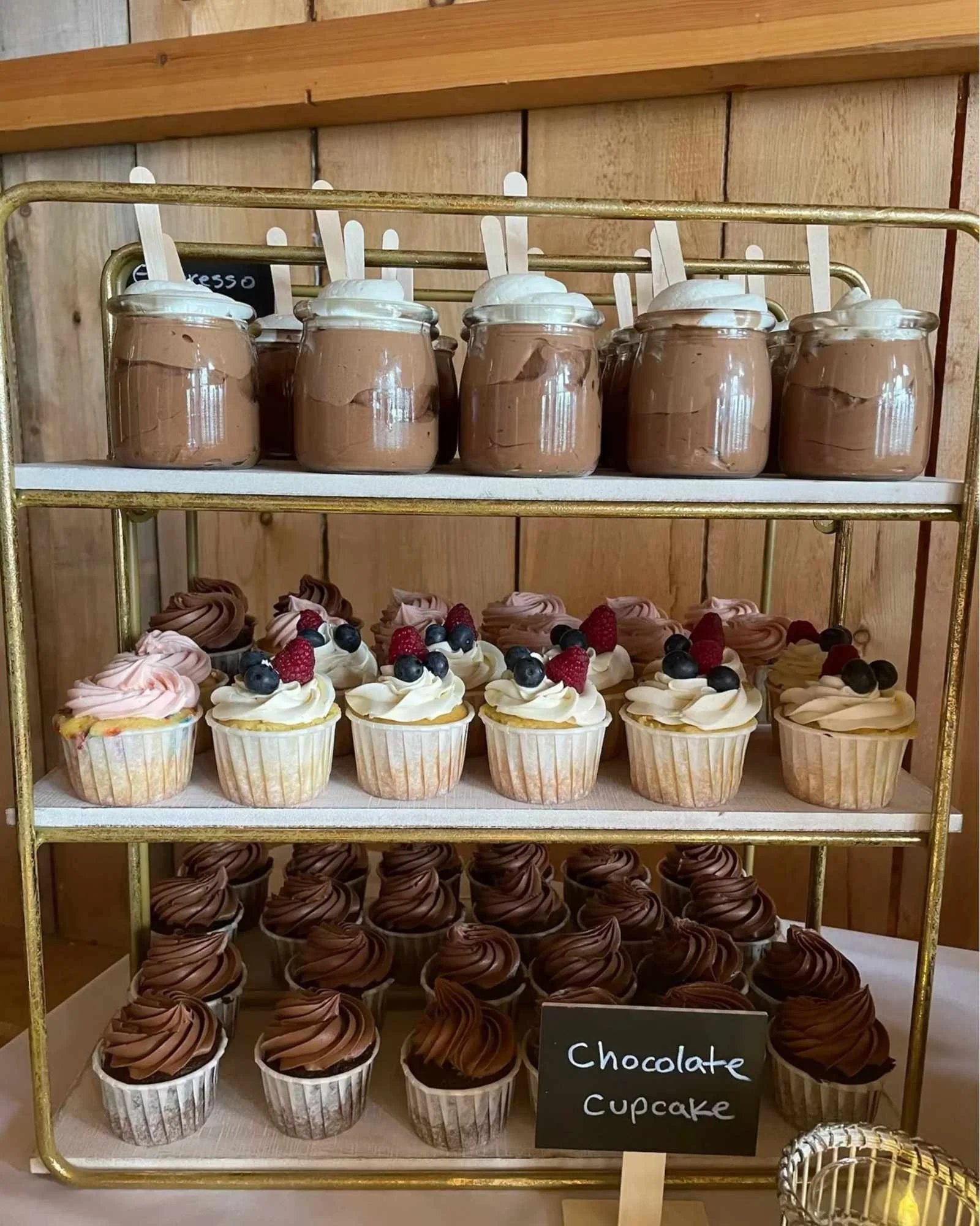 A display of chocolate cupcakes with frosting and berries, and jars of chocolate mousse with whipped topping on a gold multi-tiered stand at a bakery or café.