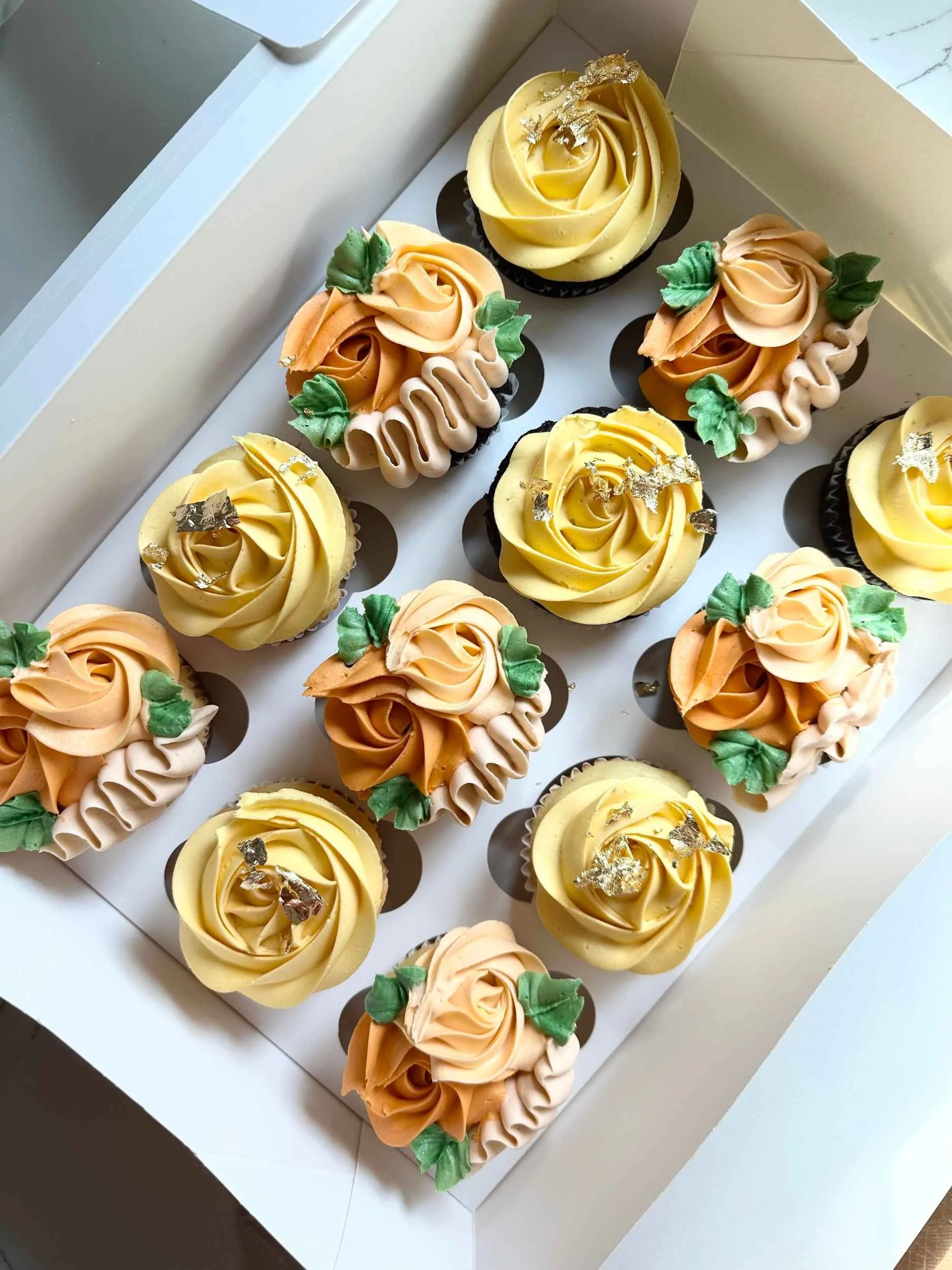 Box of assorted cupcakes decorated with yellow and orange frosting, green frosting leaves, and silver edible accents.