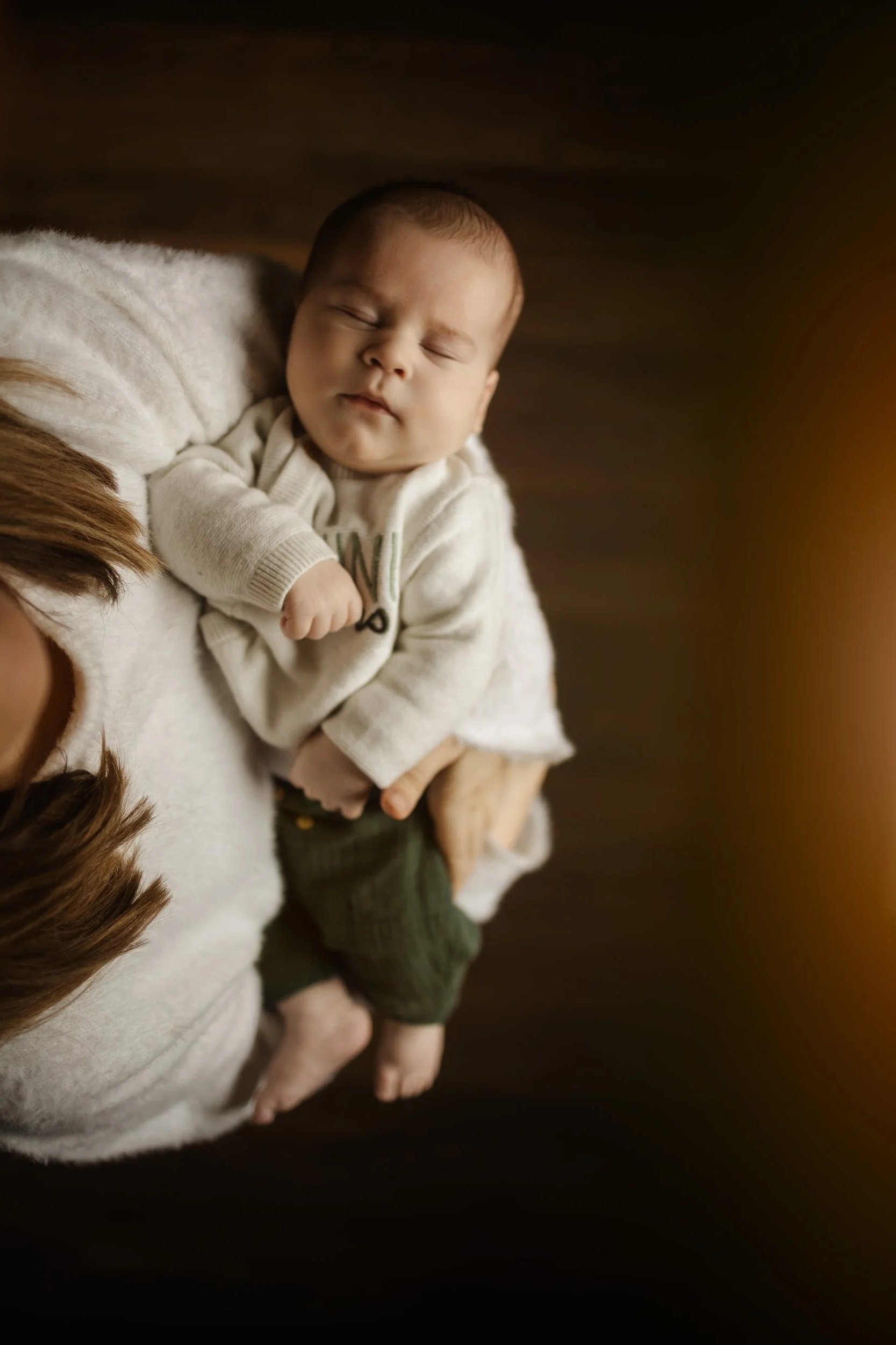 Un bébé dormant allongé sur une personne, avec des mains posées doucement sur lui, dans un environnement chaud et sombre.