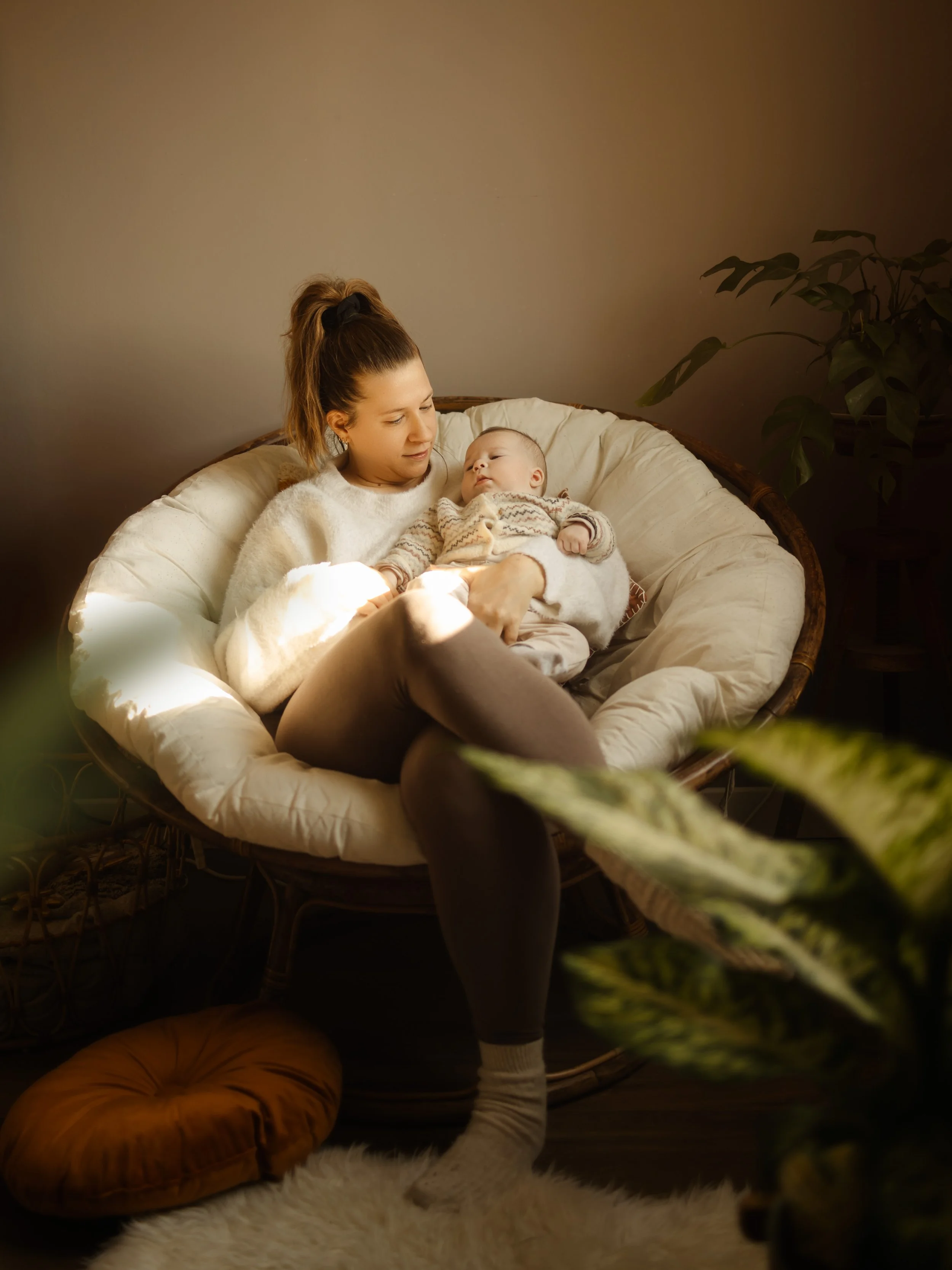 Une femme assise dans un fauteuil en rotin, tenant un bébé sur ses genoux, dans une pièce chaleureuse. La femme regarde le bébé, qui est endormi ou regarde doucement.
