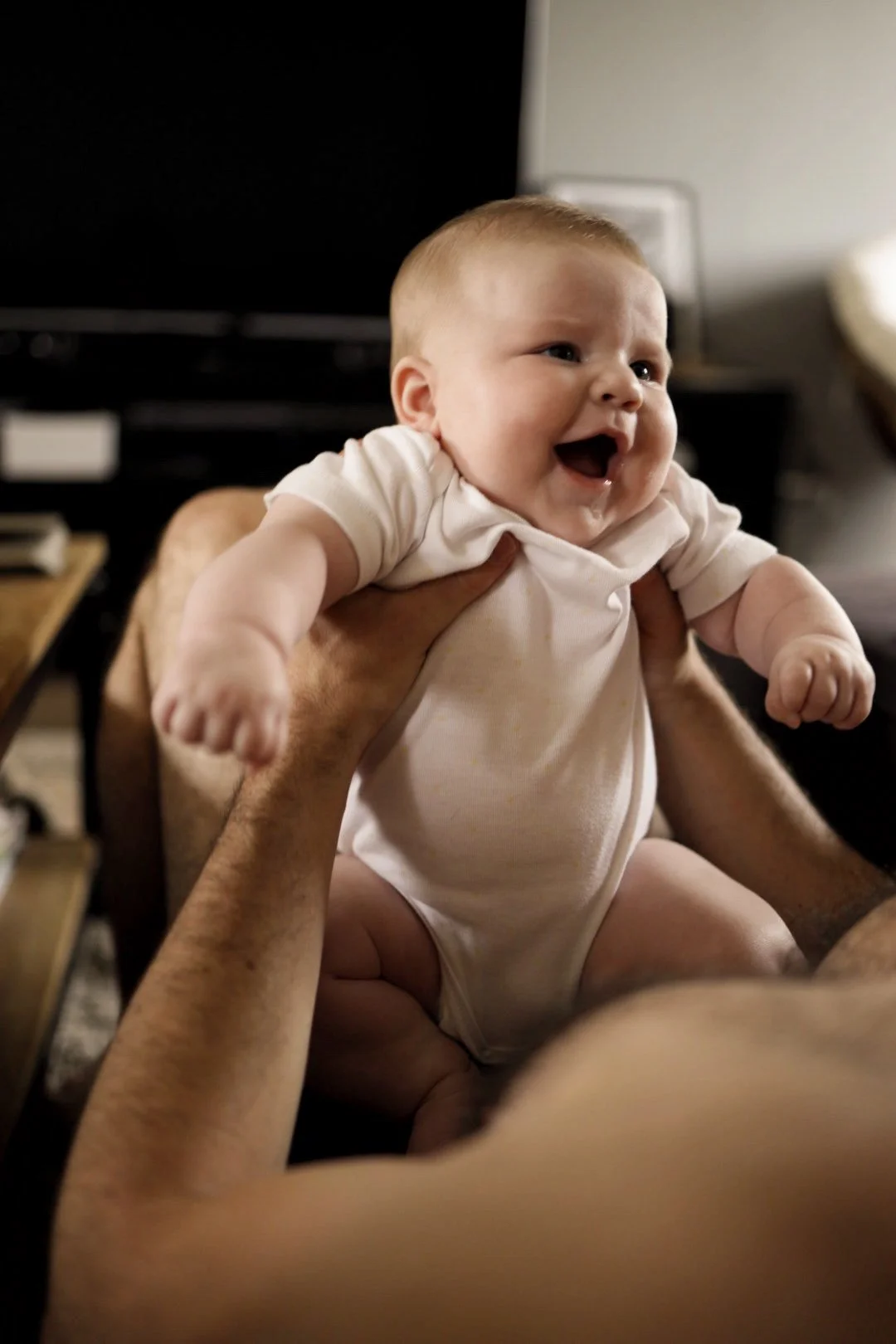 Un bébé souriant enroulé dans les bras d'une personne. L'enfant porte un body blanc et semble heureux.