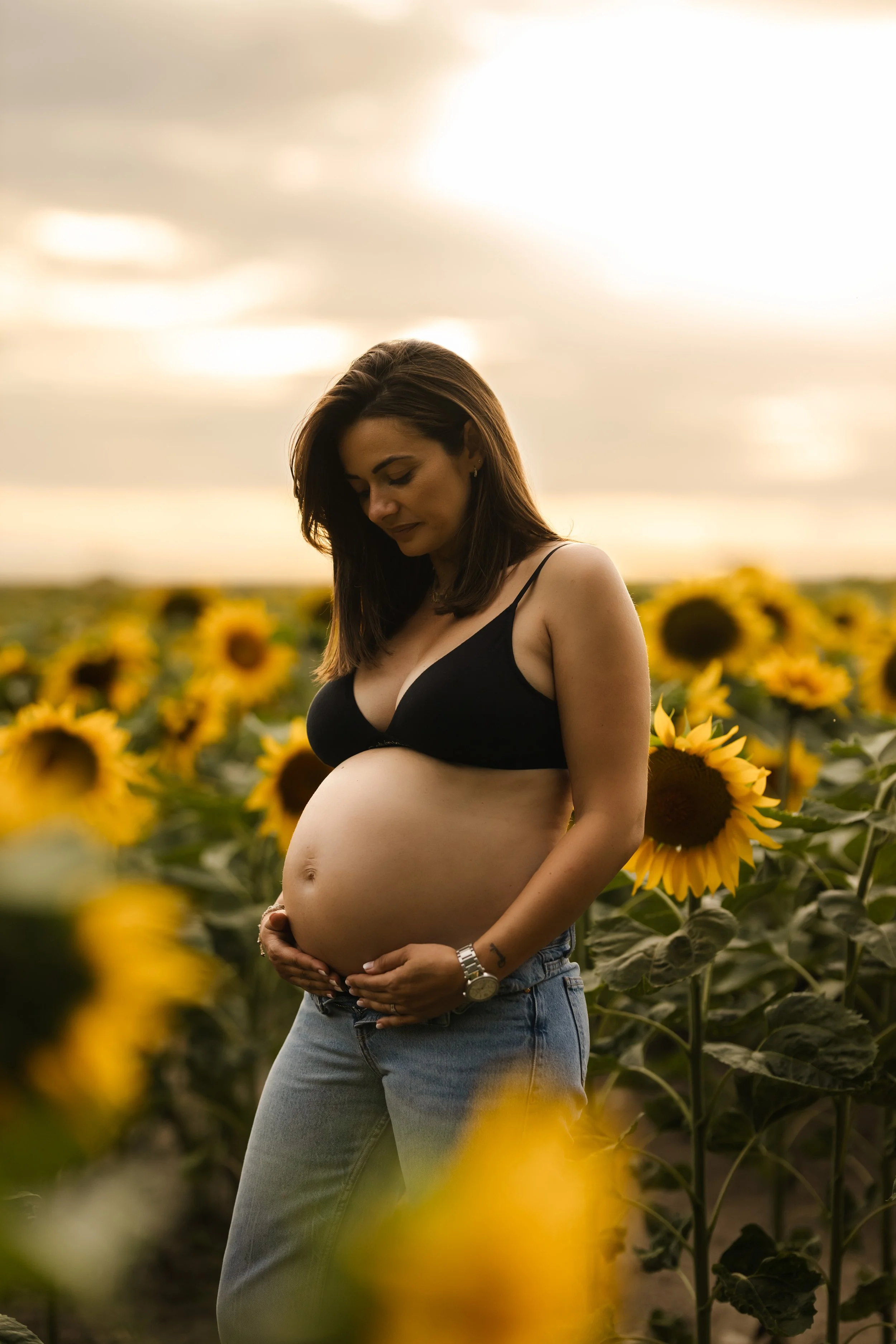 Une femme enceinte portant un haut noir et un jean, se tenant le ventre, dans un champ de tournesols sous un ciel nuageux au coucher du soleil.