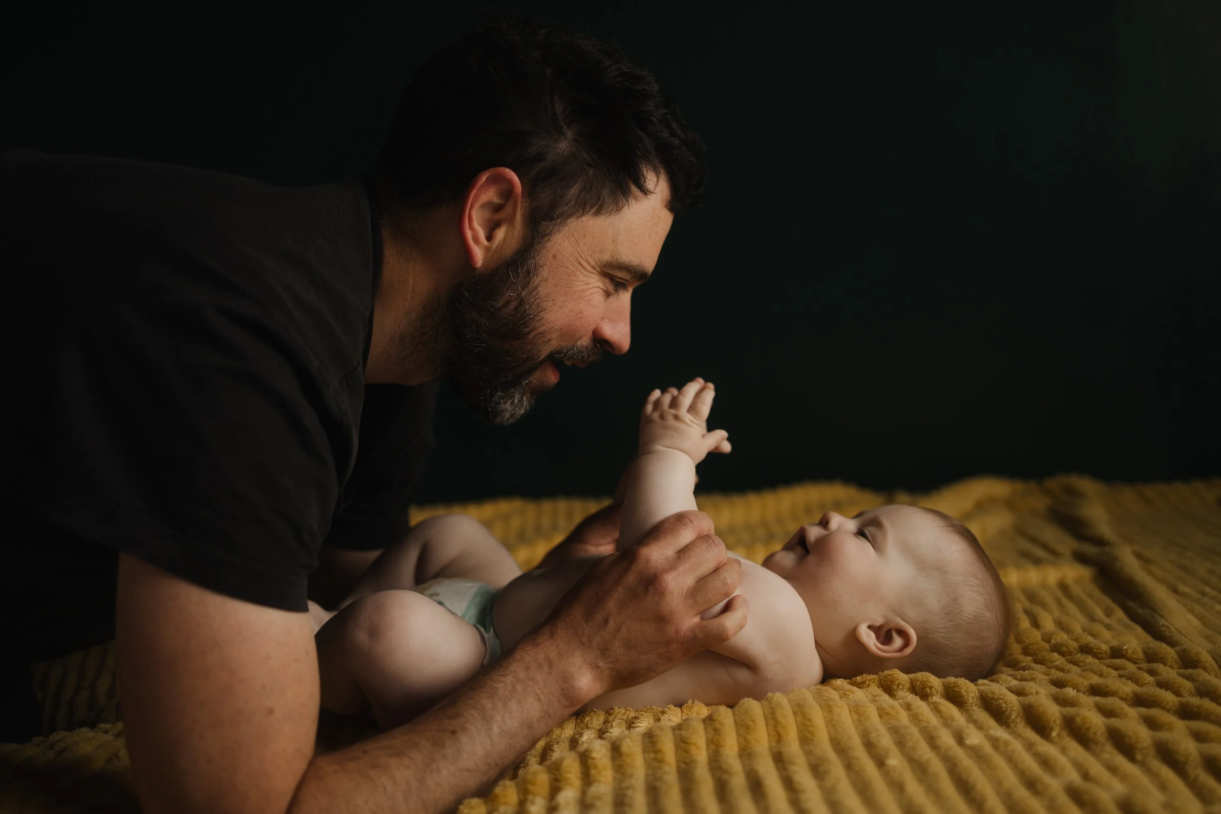 Un homme adulte joue avec un bébé allongé sur un tapis jaune, souriant et regardant le bébé tout en tenant sa main.