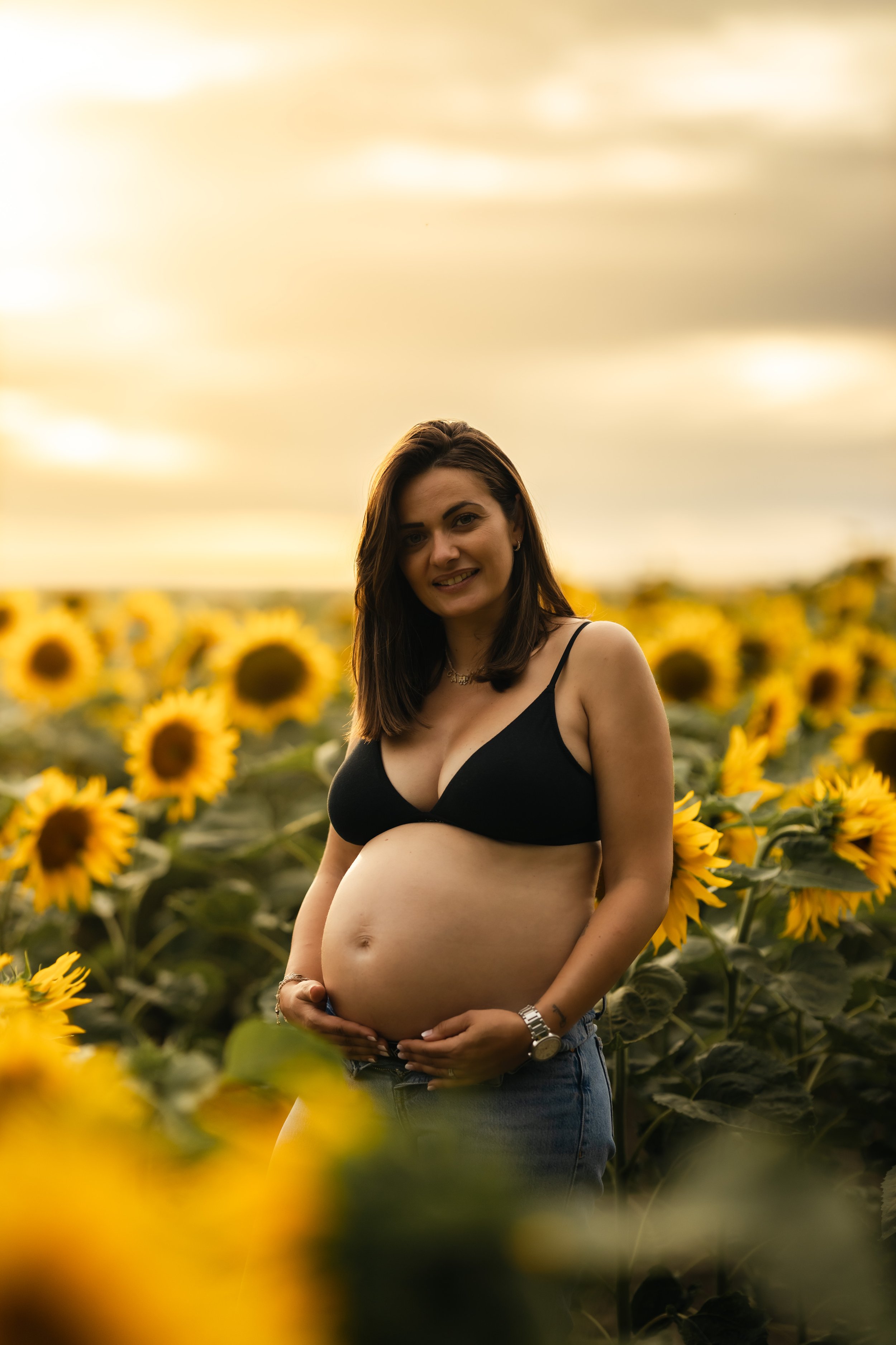 Femme enceinte souriante portant un haut noir dans un champ de tournesols au coucher du soleil.