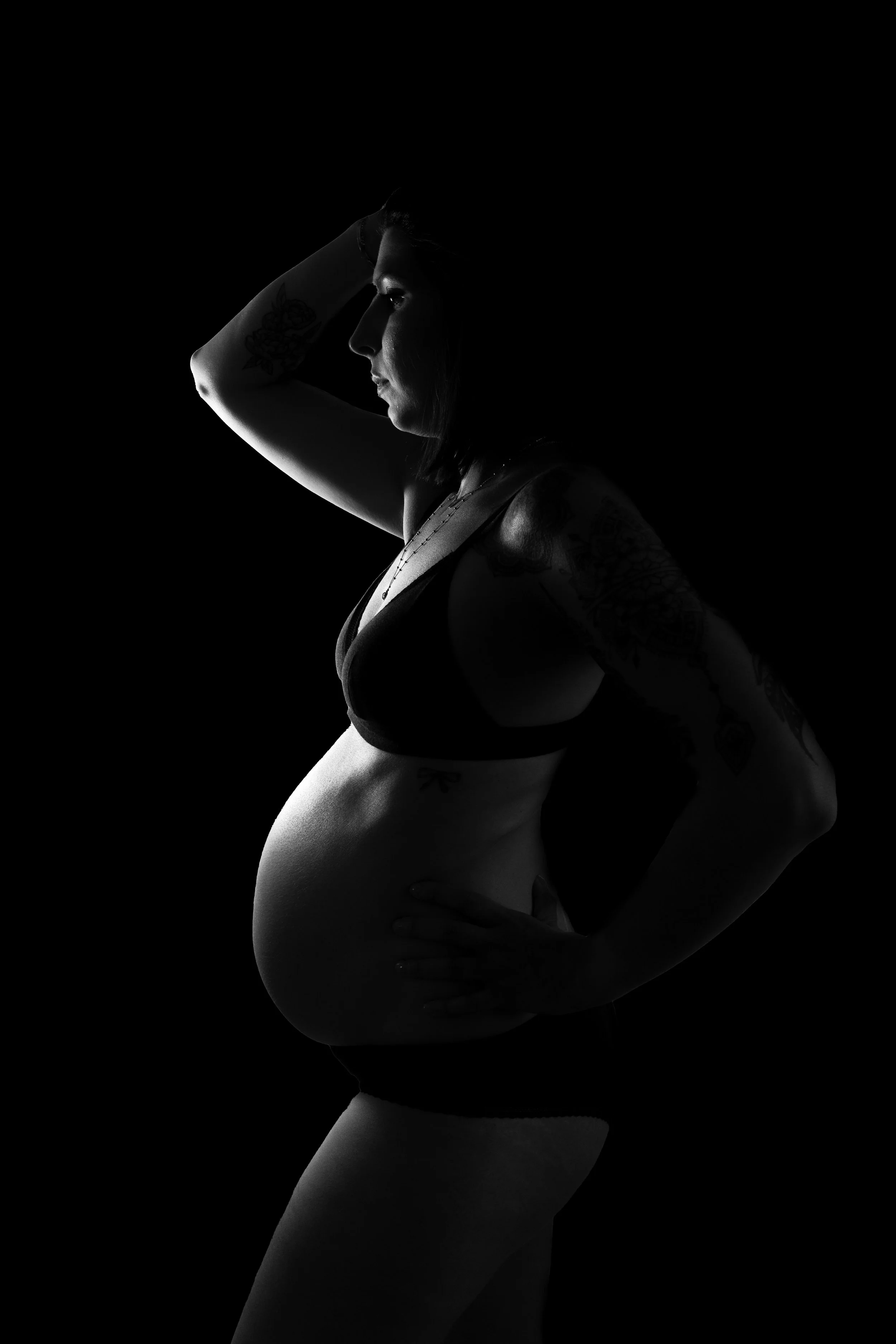 Silhouette en noir et blanc d'une femme enceinte, portant un soutien-gorge noir et des sous-vêtements, avec un bras sur la taille et l'autre derrière la tête, mettant en valeur son ventre rond, avec des tatouages sur le bras