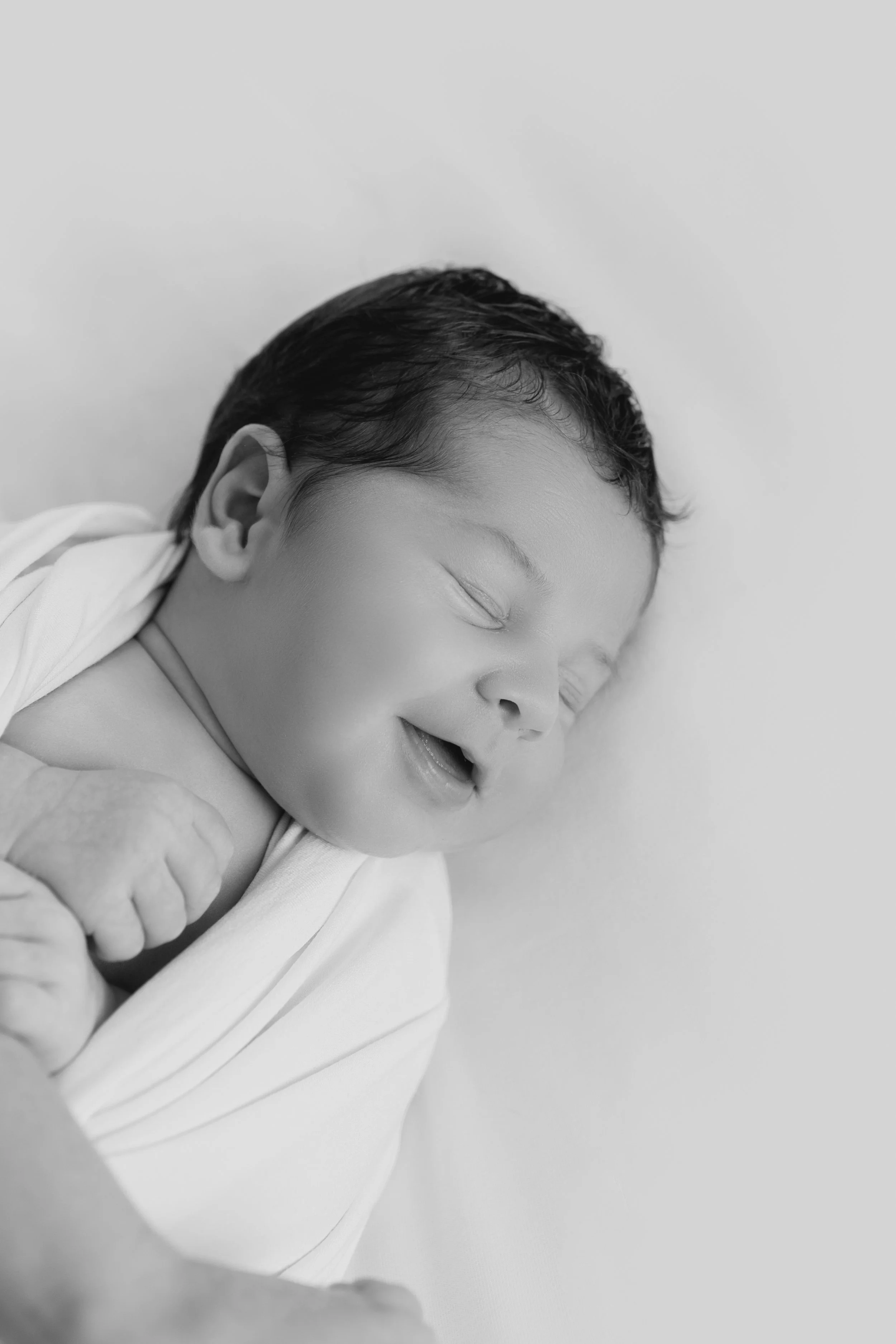Un bébé souriant endormi, en noir et blanc, couché sur un lit ou une surface douce.